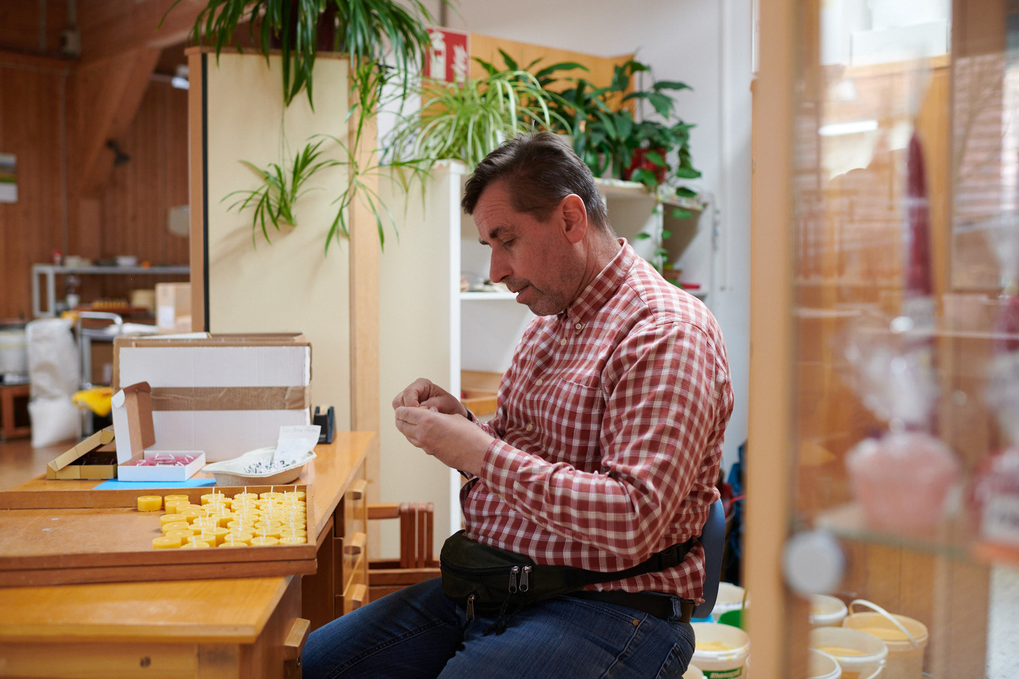 Mann beim Zusammenbau von Produkten am Schreibtisch Ein Mann in kariertem Hemd und Jeans montiert kleine gelbe Teile an einem Tisch mit Pflanzen im Hintergrund.
