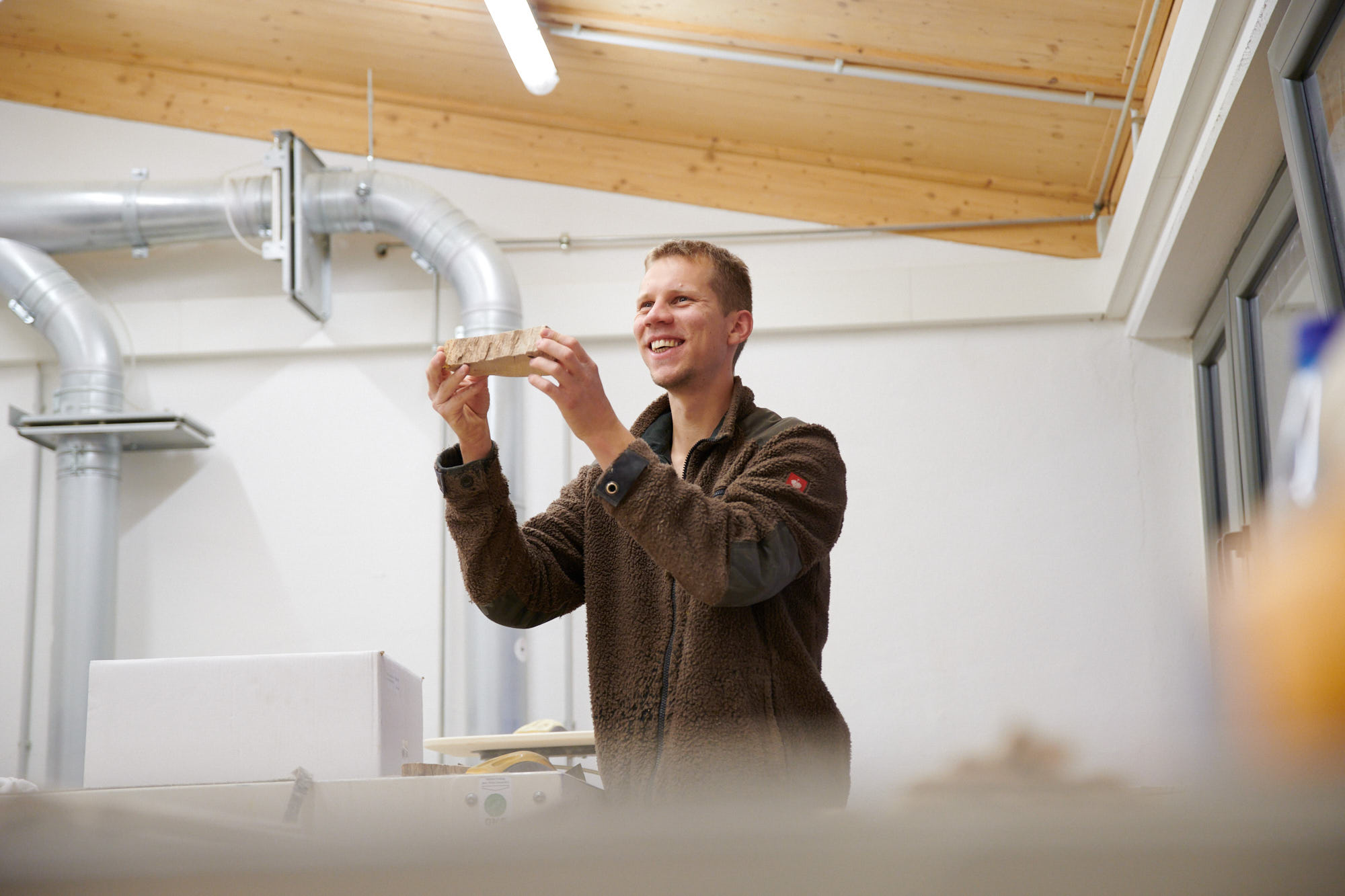 Mann in Werkstatt mit Holzstück Ein Mann in einer Werkstatt hält lächelnd ein Holzstück hoch.