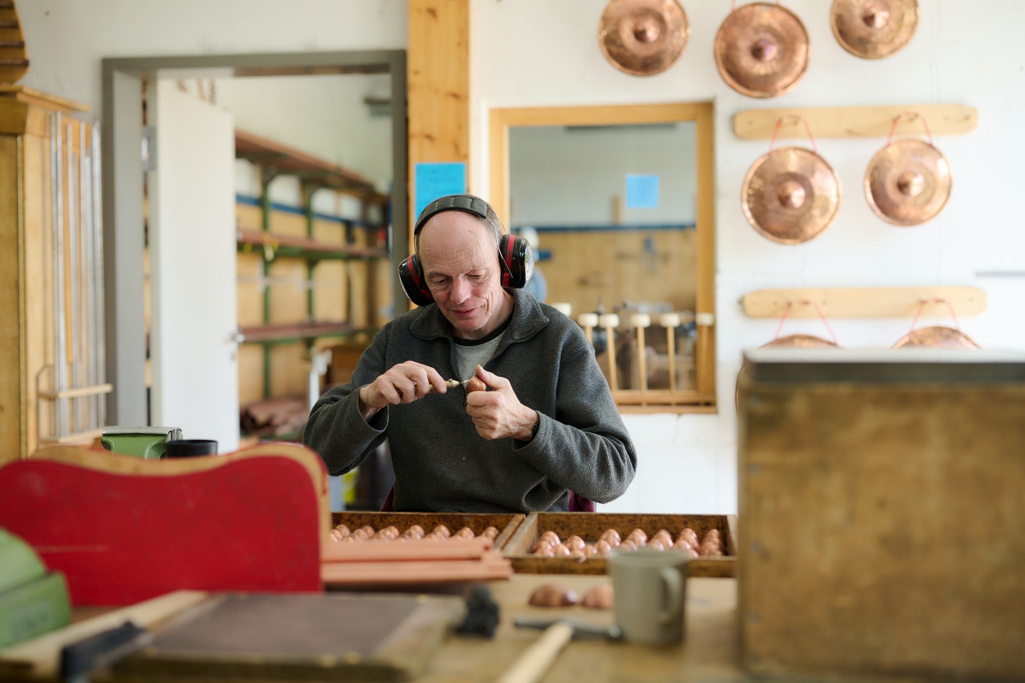 Handwerker bei der Arbeit in einer Werkstatt Ein Handwerker, der Gehörschutz trägt, arbeitet konzentriert an einem Objekt in einer Werkstatt, umgeben von Handwerkszeug und Kupfergegenständen.
