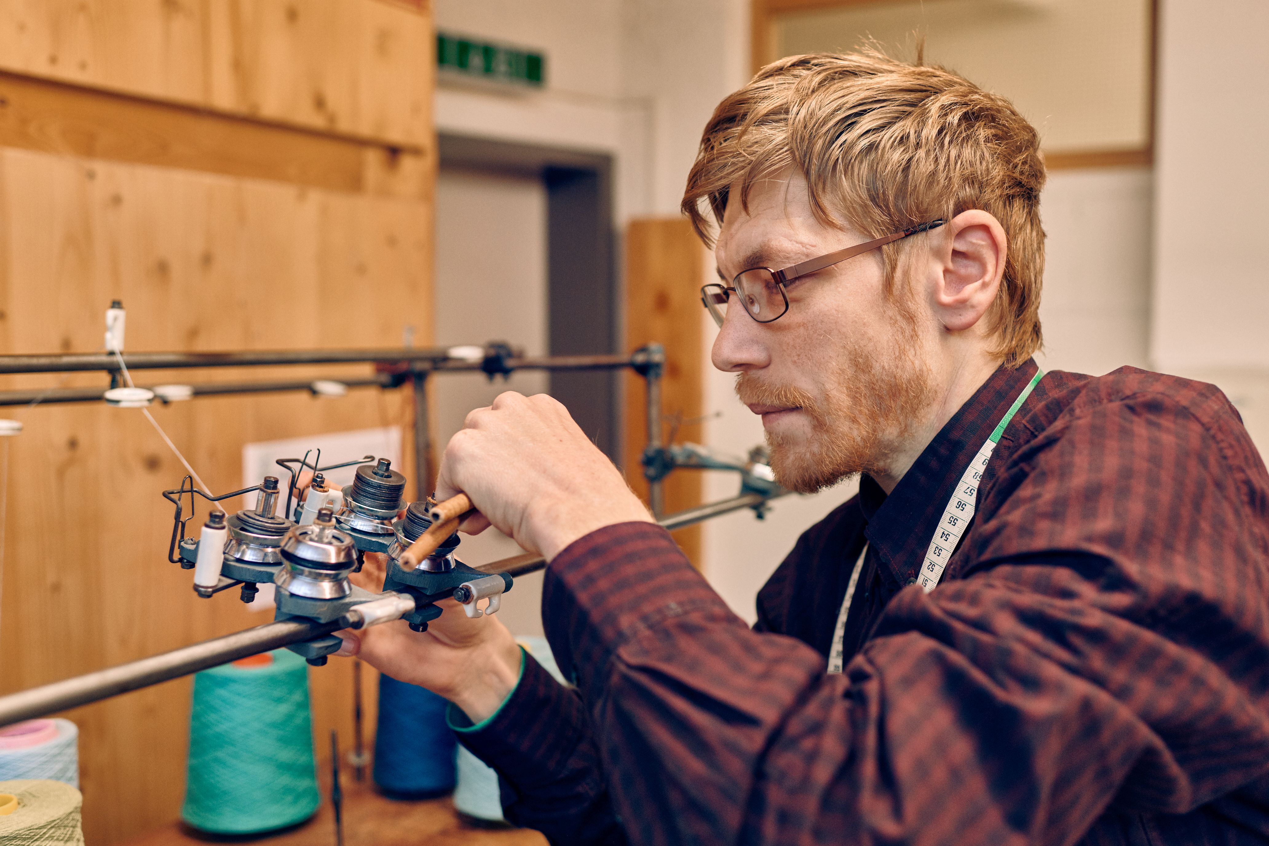 Präzise Arbeit an einer Textilmaschine Ein Mann mit Brille und kariertem Hemd arbeitet sorgfältig an einer Textilmaschine in einem Atelier.
