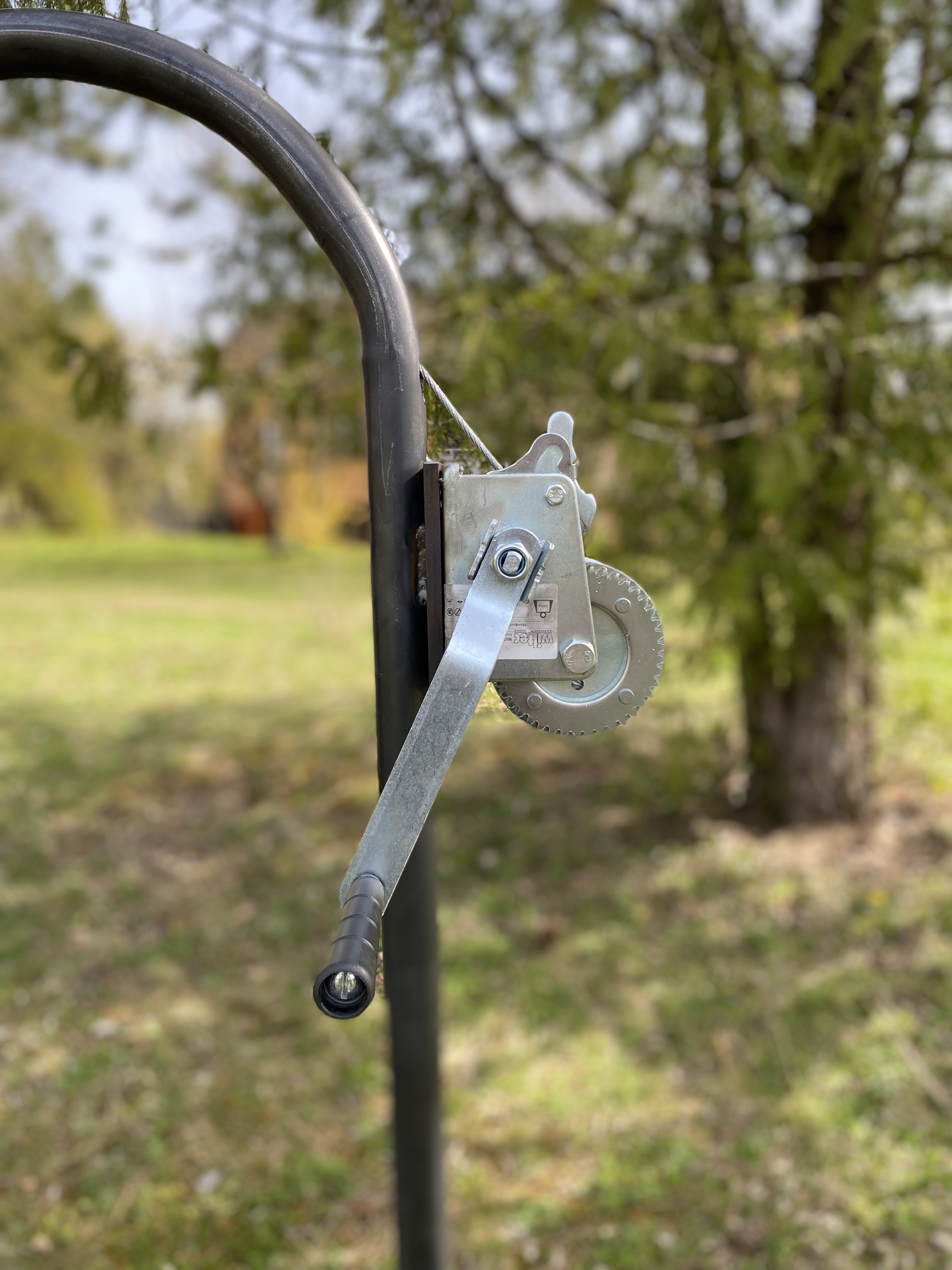 Nahaufnahme einer montierten Handseilwinde mit Kurbel an einem schwarzen Metallrohrgestell im Garten