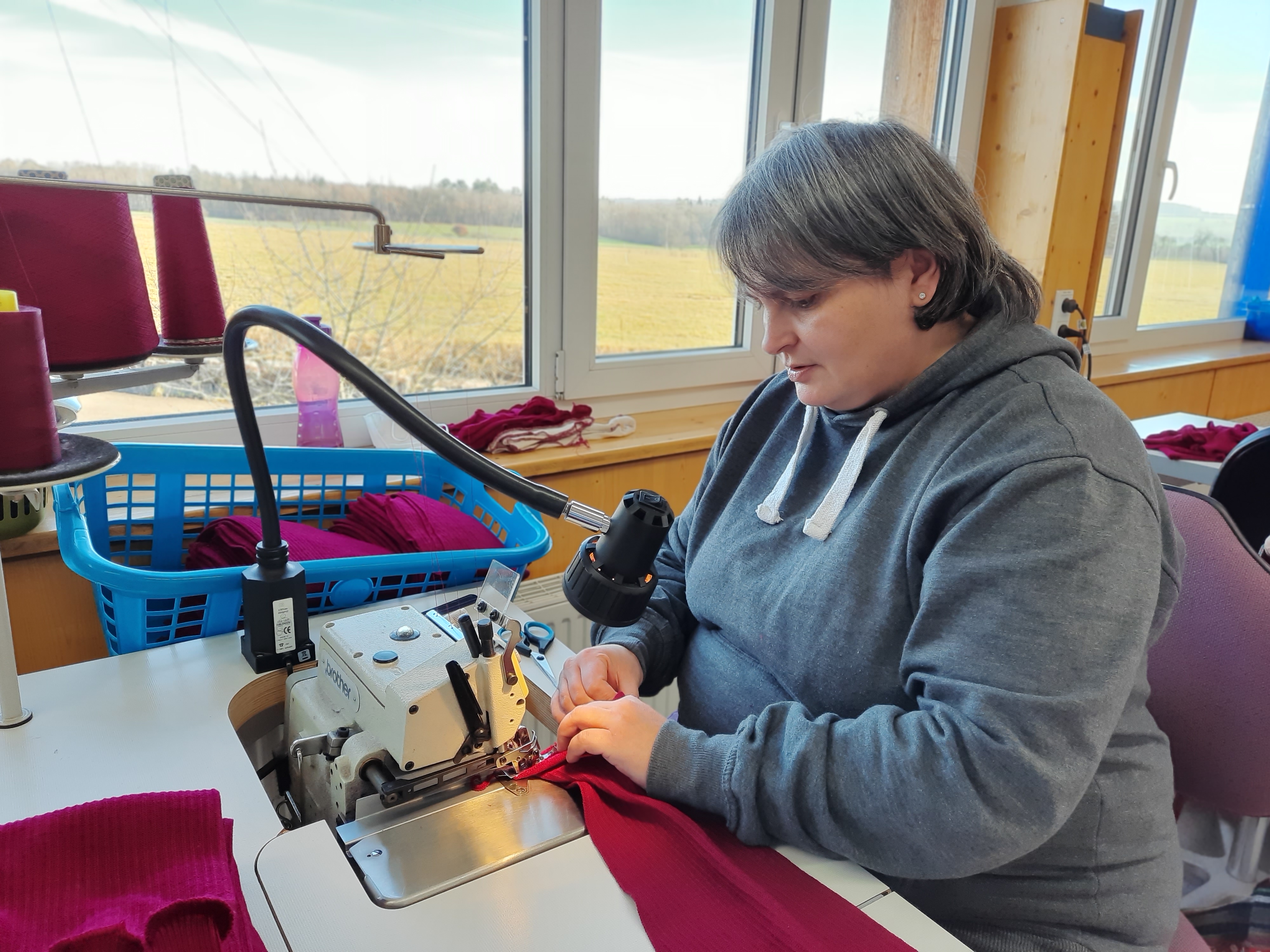 Frau näht mit einer Overlock-Maschine in einem hellen Raum Person in grauem Hoodie näht roten Stoff mit einer Overlock-Maschine neben einem Fenster mit Blick auf eine Landschaft