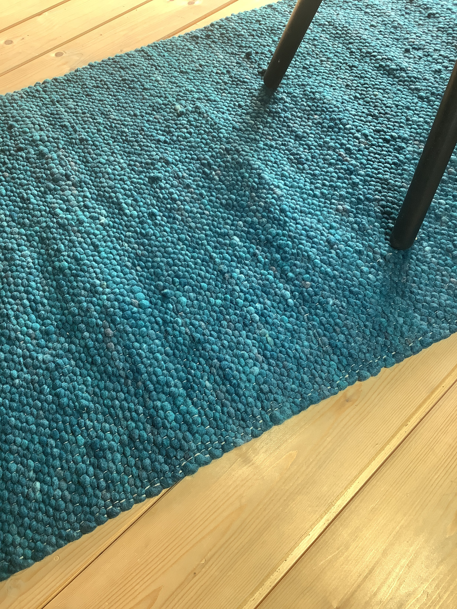 Detailansicht eines türkisfarbenen Läuferteppichs aus Wollkugeln auf einem hellen Holzboden mit Stuhlbeinen