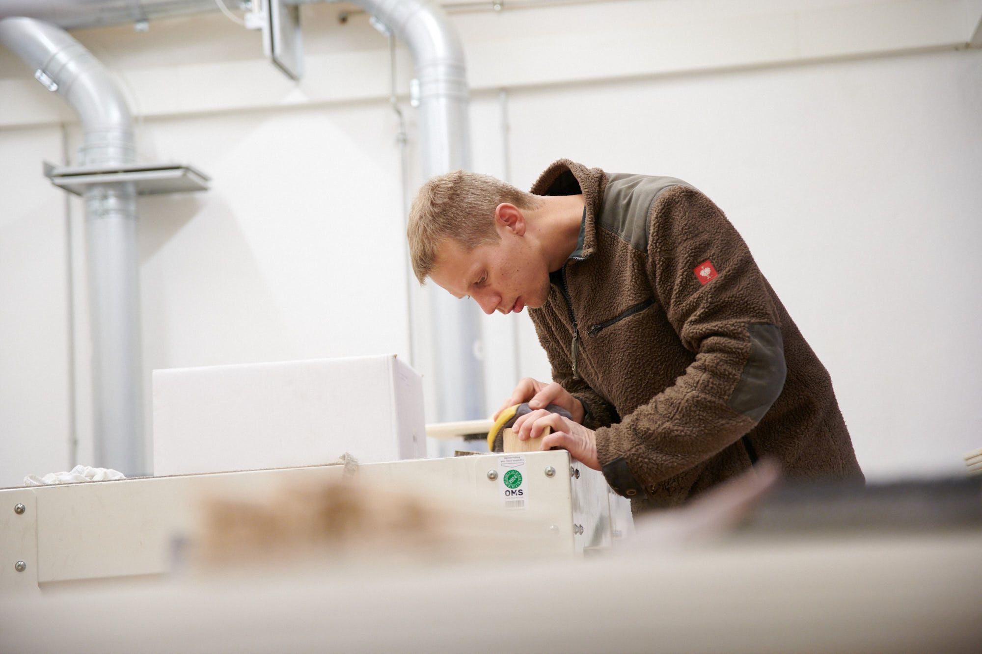 Ein Mann in einer braunen Fleecejacke arbeitet konzentriert in einer Fabrikumgebung an einer Maschine.