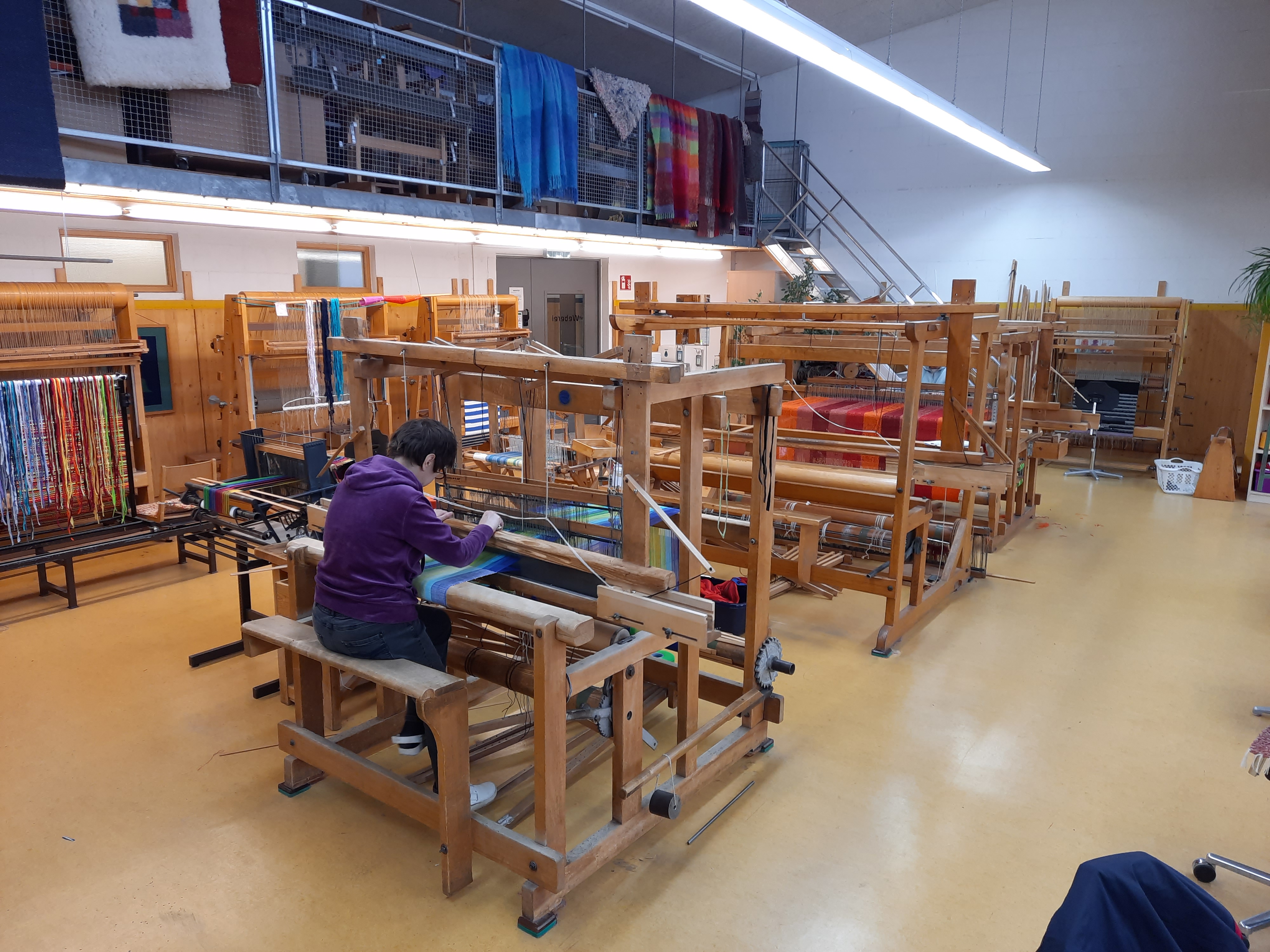 Frau am Webstuhl in einer Weberei Eine Person arbeitet an einem großen Webstuhl in einer traditionellen Weberei, umgeben von bunten Stoffen.