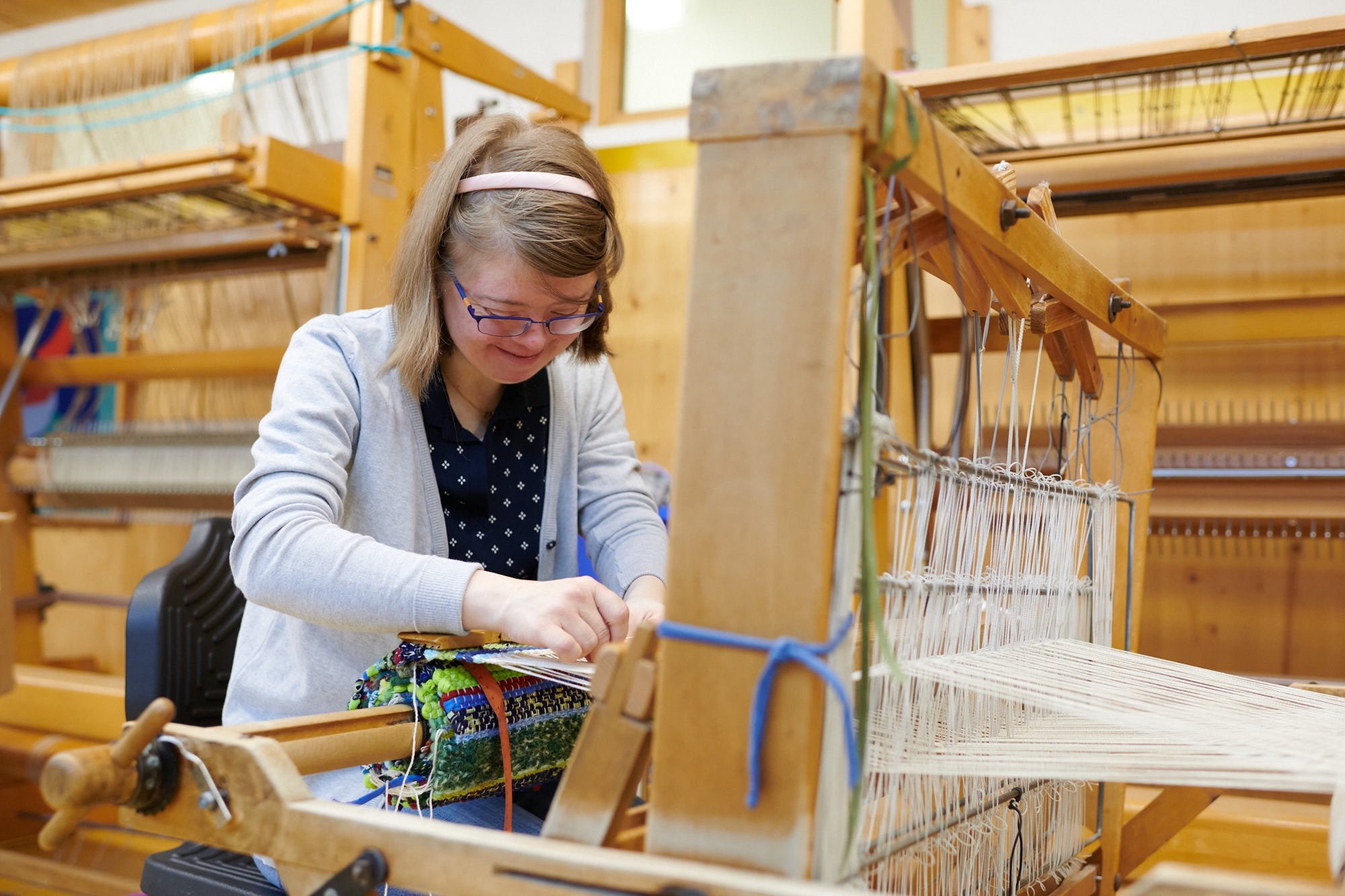 Frau beim Weben an einem Holzwebstuhl Eine Frau mit blauer Brille webt konzentriert an einem traditionellen Holzwebstuhl in einer Werkstatt.