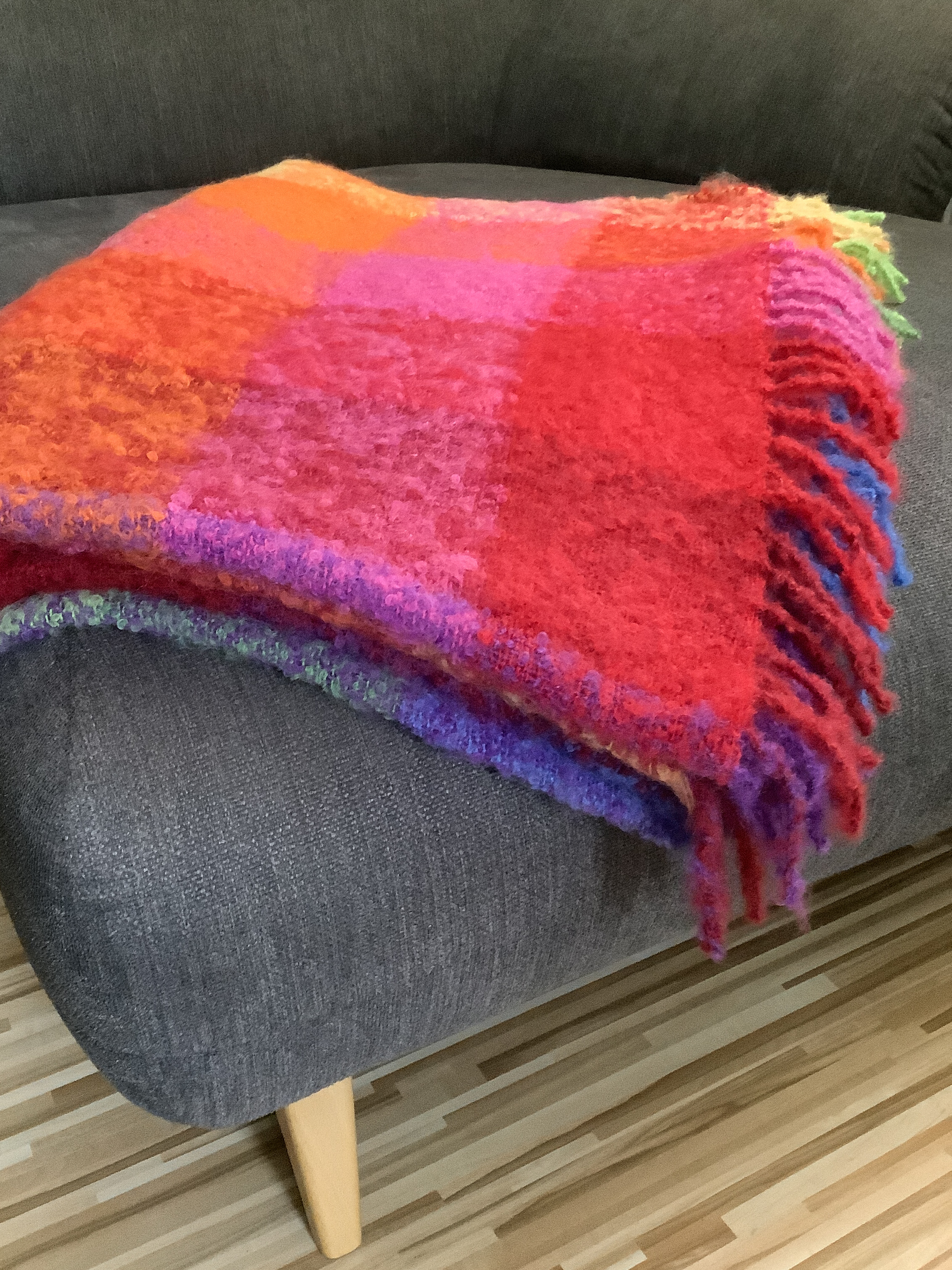 Farbenfrohe Decke aus Wolle auf grauem Sofa Bunte Patchworkdecke aus Wolle mit roten, orangefarbenen und lilafarbenen Quadraten, liegend auf einer Sofalehne aus grauem Stoff