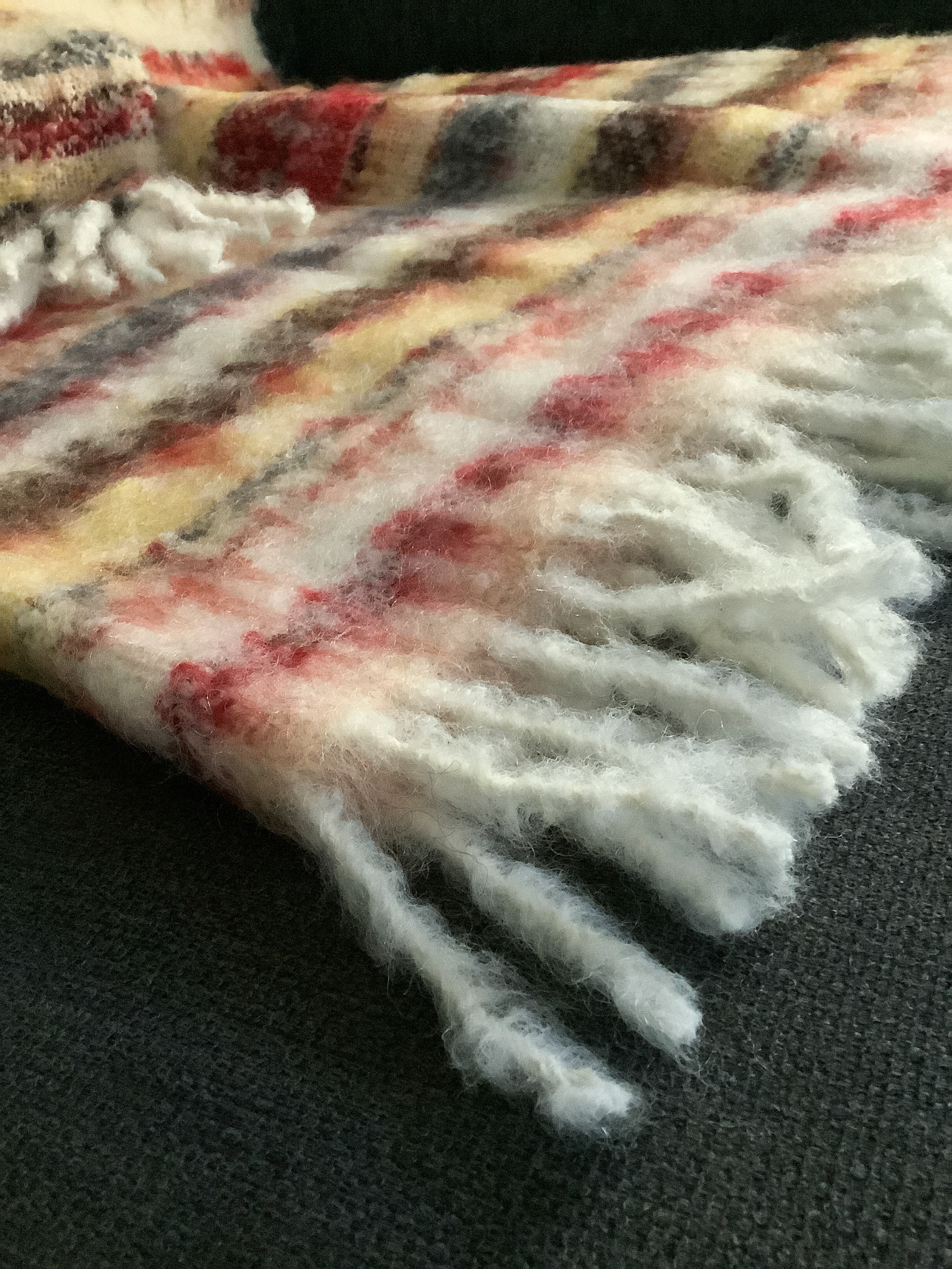 Weiche, bunte Kuscheldecke mit Fransen auf einem Sofa Nahaufnahme einer gestrickten Decke in bunten Farben mit Fransen auf einem dunklen Sofa