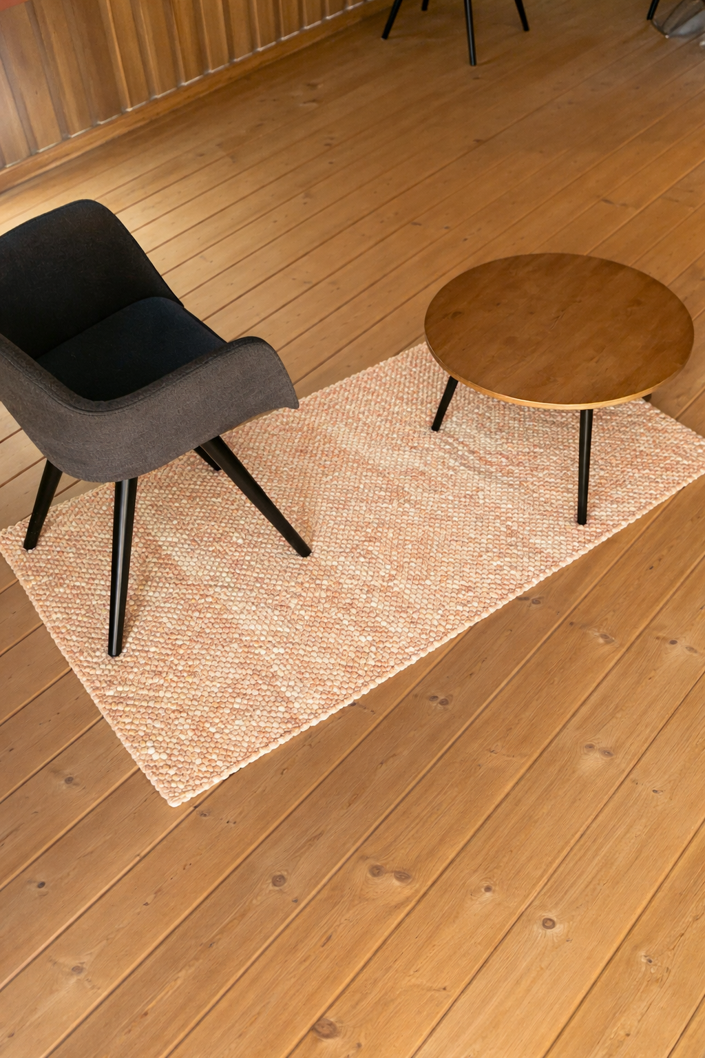 Heller rechteckiger Teppich unter rundem Holztisch und dunkelgrauem Polsterstuhl auf warmem Holzboden in modernem Wohnbereich