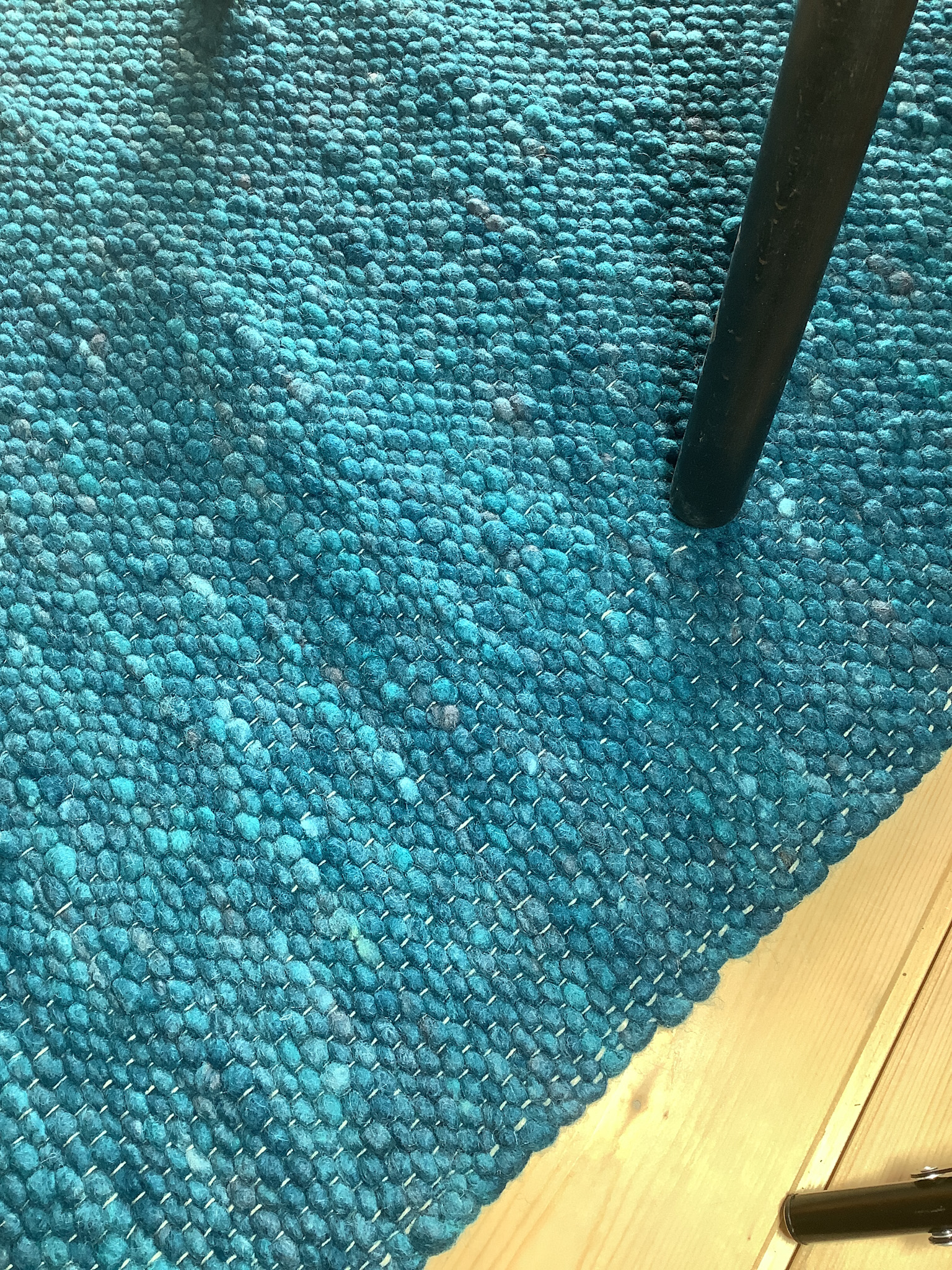 Nahaufnahme eines türkisblauen, grob strukturierten Wollteppichs auf hellem Holzboden mit Tischbein
