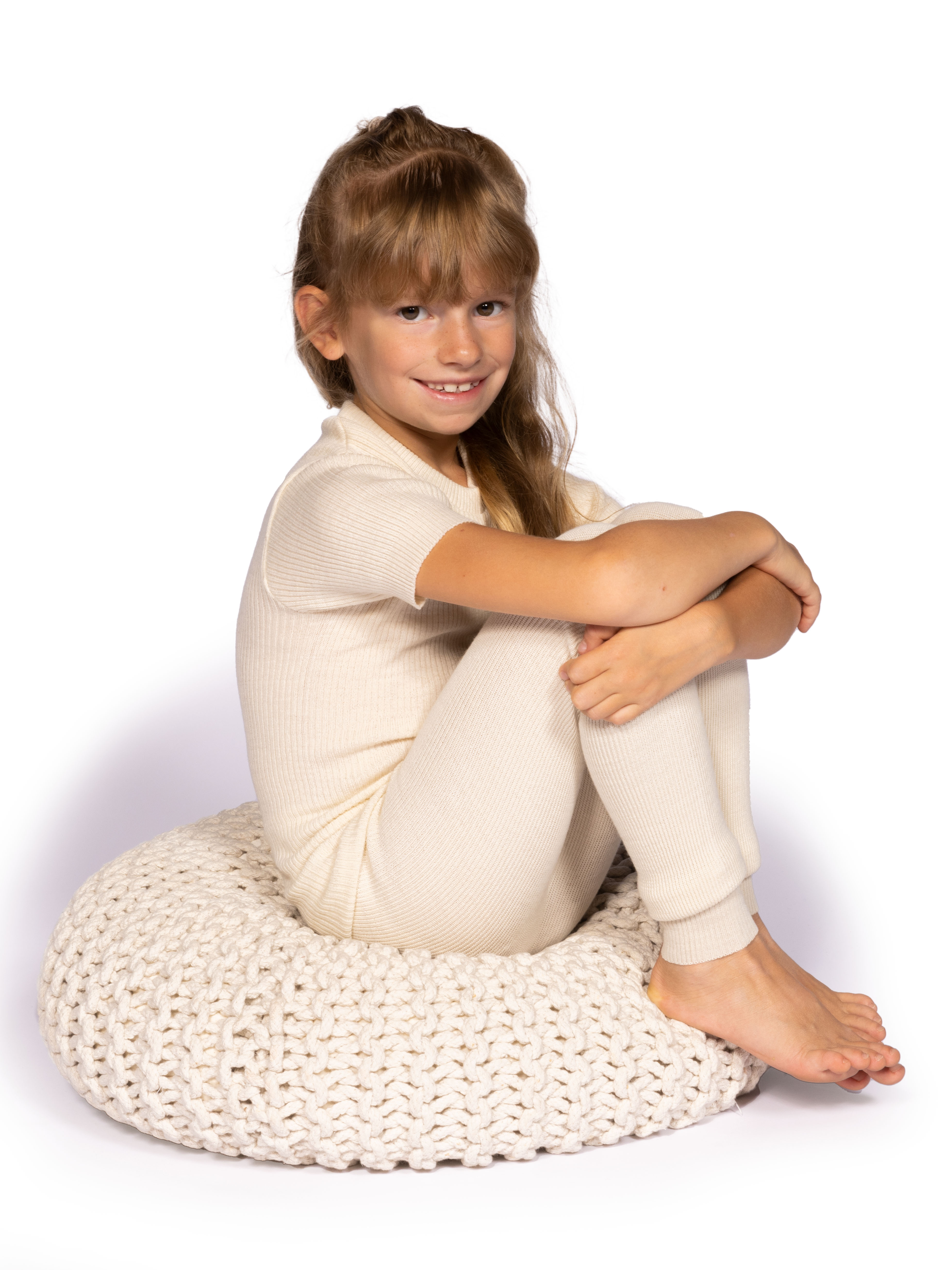 Mädchen in cremeweißer Kleidung auf gestricktem Kissen Ein lächelndes Mädchen sitzt im Schneidersitz auf einem cremefarbenen gestrickten Kissen und trägt passende, warme Kleidung.