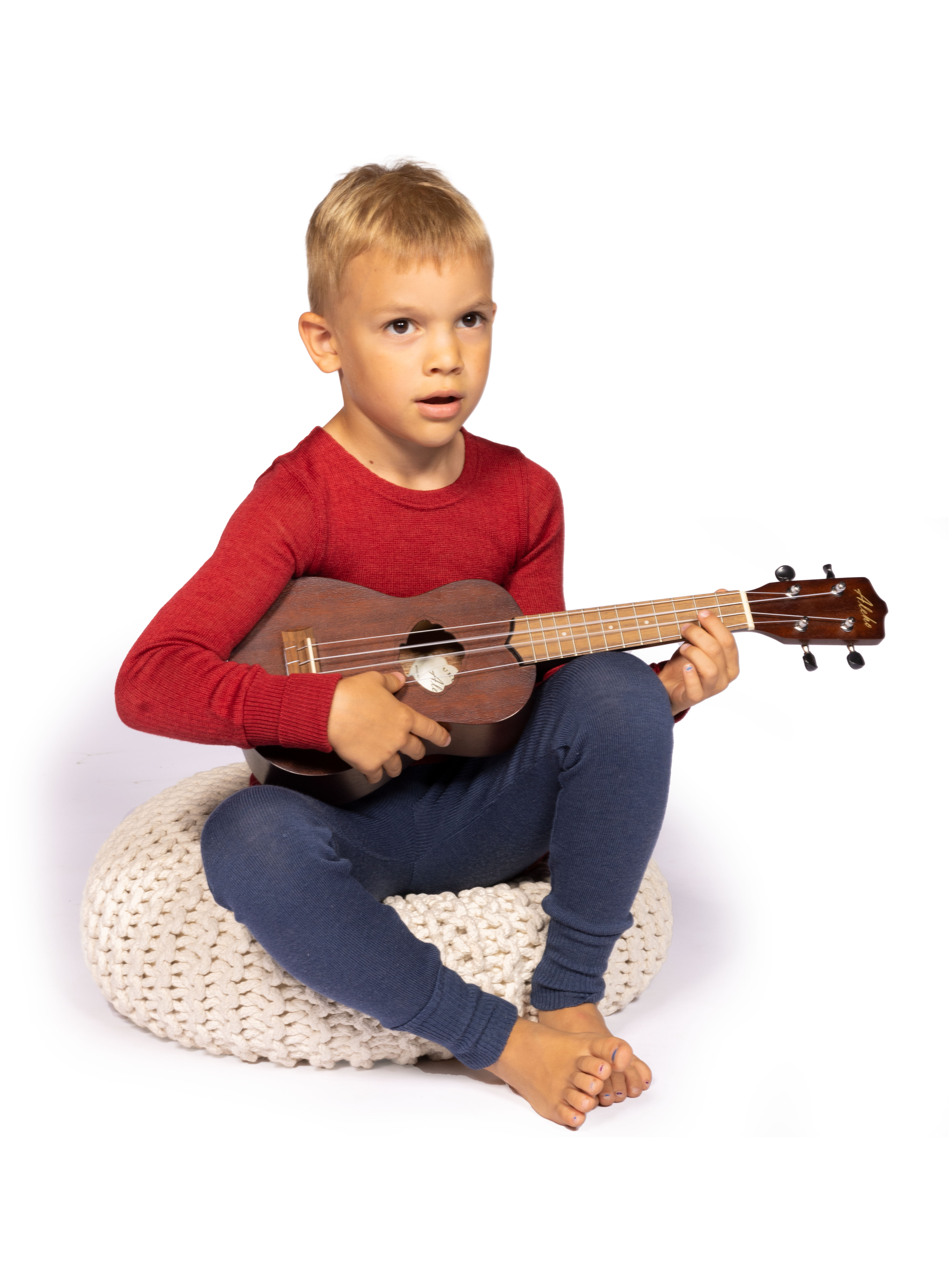 Kind spielt Ukulele auf einem Strickhocker Junge mit rotem Pullover und blauer Hose spielt Ukulele, sitzend auf weißem Strickhocker