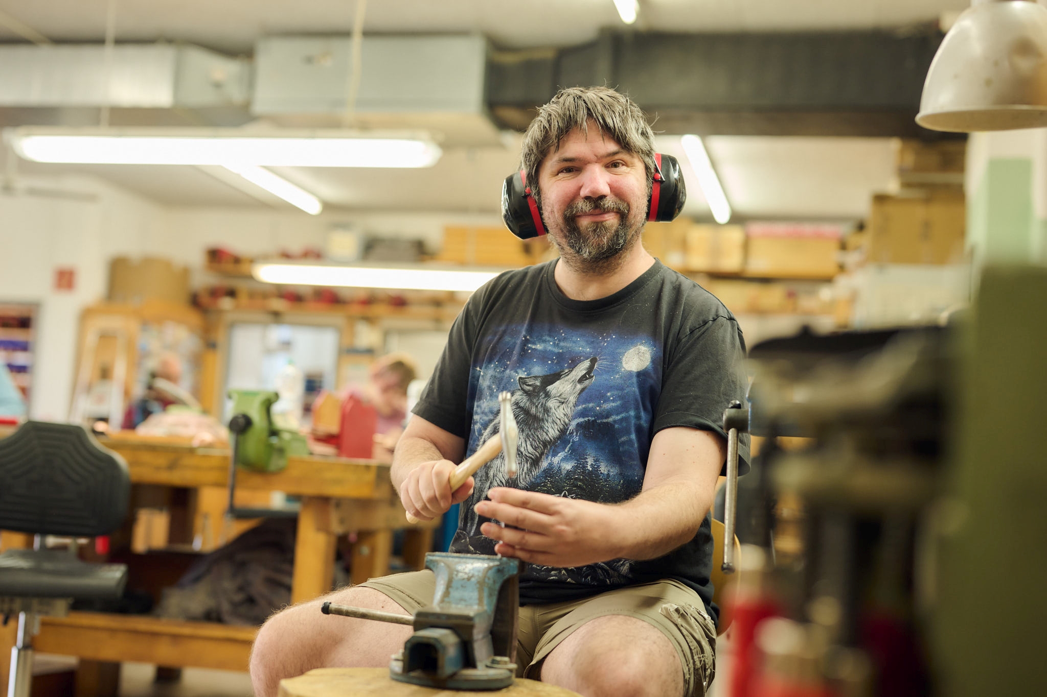 Handwerker bei der Arbeit in einer Werkstatt mit Gehörschutz Ein Mann mit Gehörschutz arbeitet in einer Werkstatt und benutzt einen Hammer, während andere Personen im Hintergrund zu sehen sind.