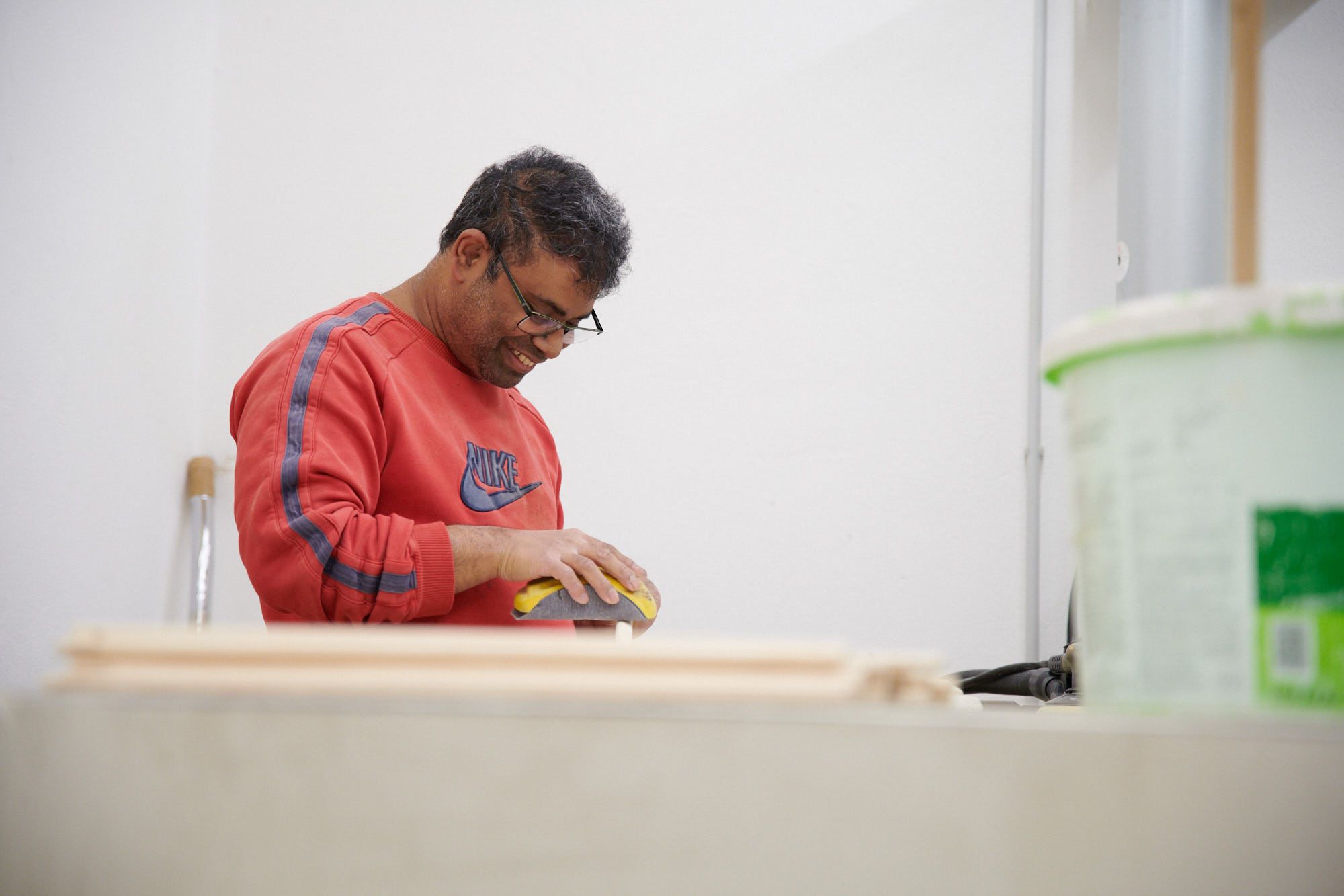 Ein Mann in einem roten Pullover schleift ein Holzstück in einer Werkstatt, im Vordergrund ein Farbeimer