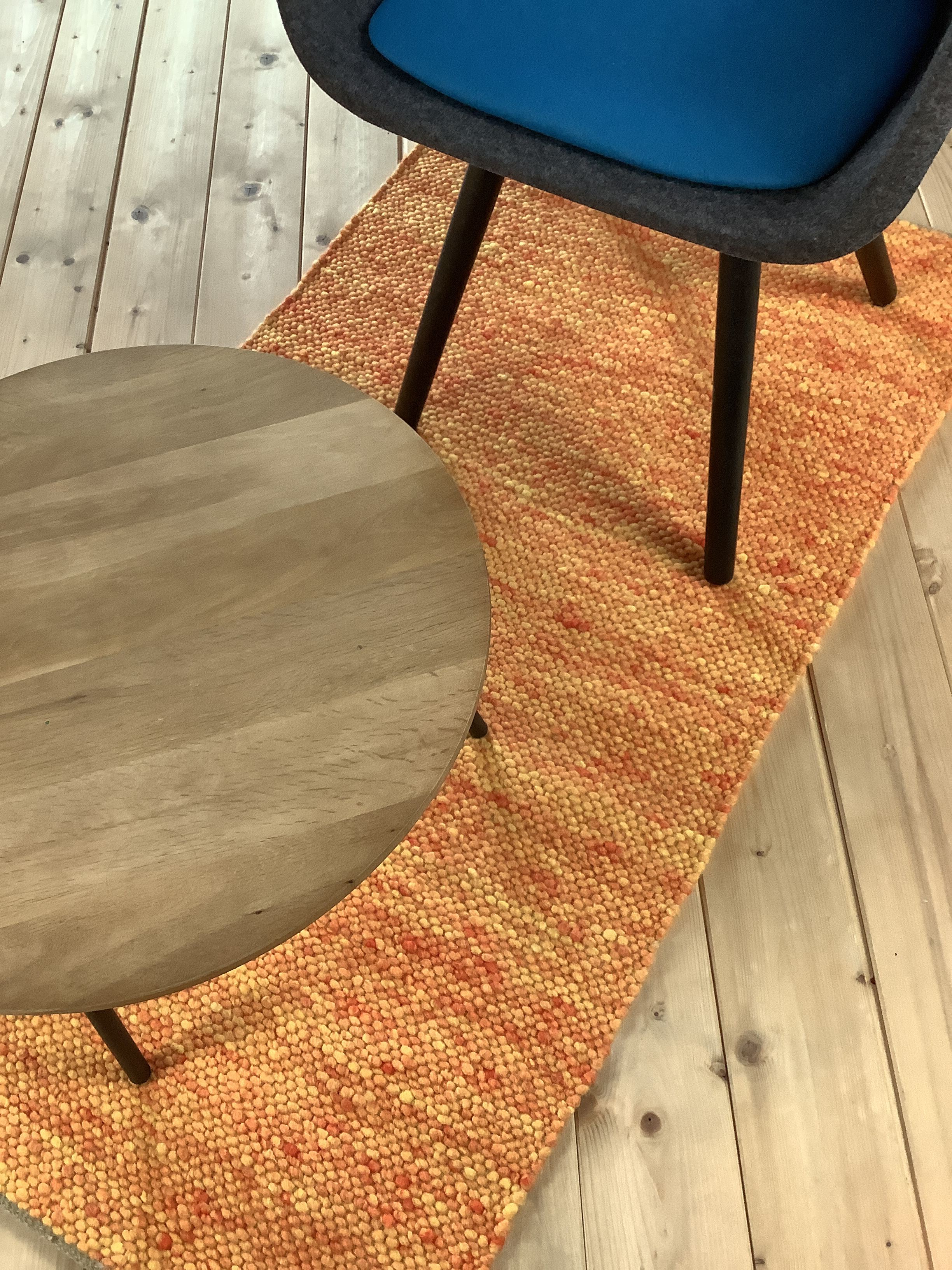 Designer-Stuhl und runder Holztisch auf buntem Teppich Ein grauer Stuhl mit blauer Sitzfläche steht neben einem runden Holztisch auf einem orangefarbenen Teppich auf Holzboden.