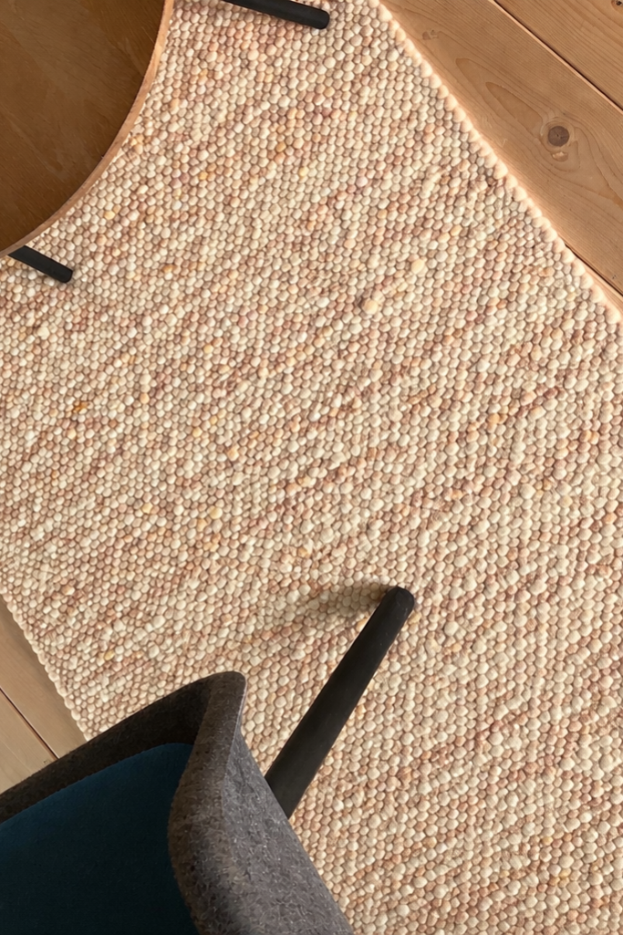 Detailansicht eines creme-beigen, handgewebten Wollteppichs mit strukturierter Oberfläche auf Holzboden, teils mit Stuhlbeinen