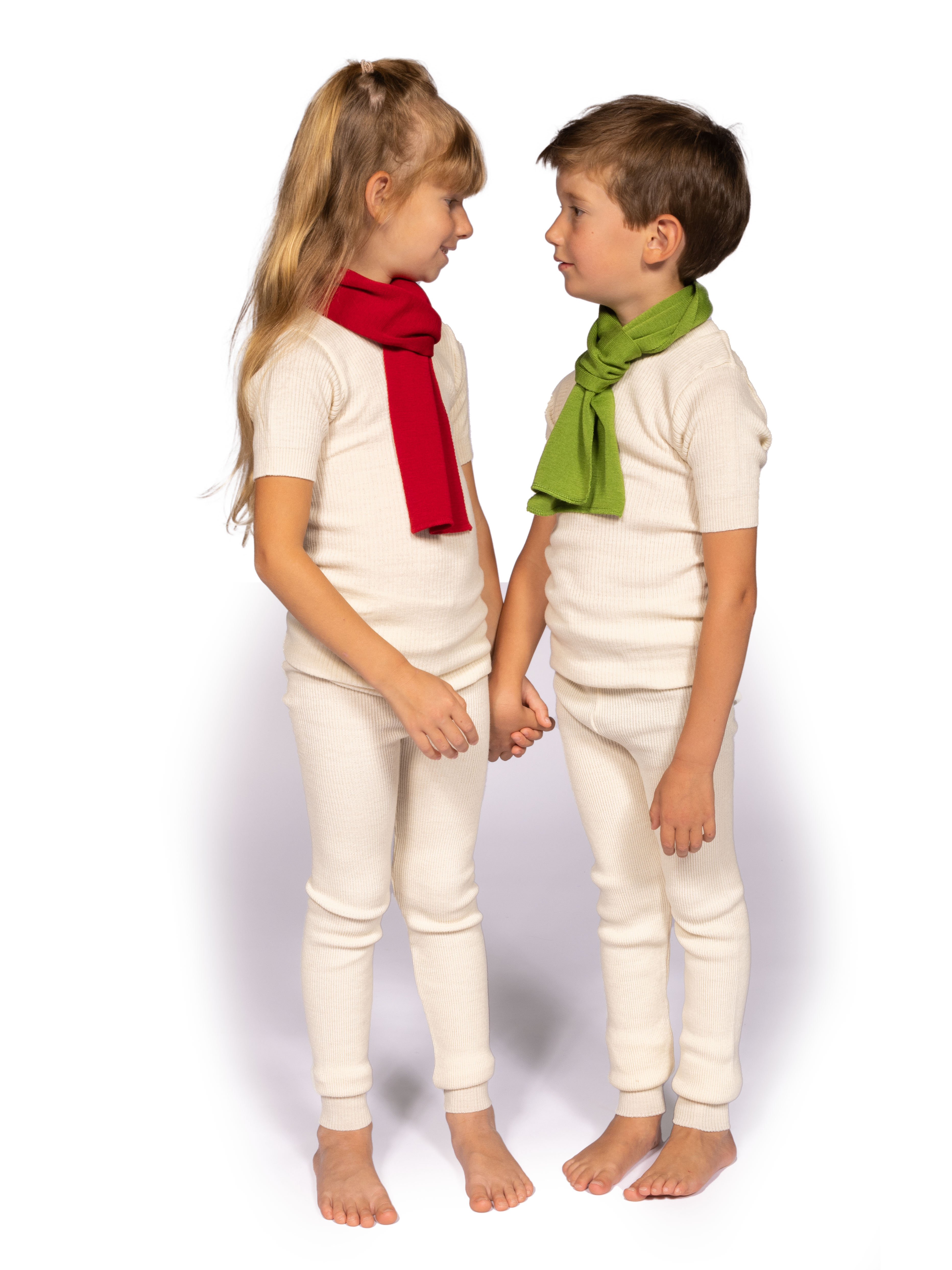 Kinder in weißer Wollunterwäsche mit bunten Schals Zwei Kinder in weißer Wollunterwäsche, ein Mädchen mit rotem Schal und ein Junge mit grünem Schal, halten sich an den Händen