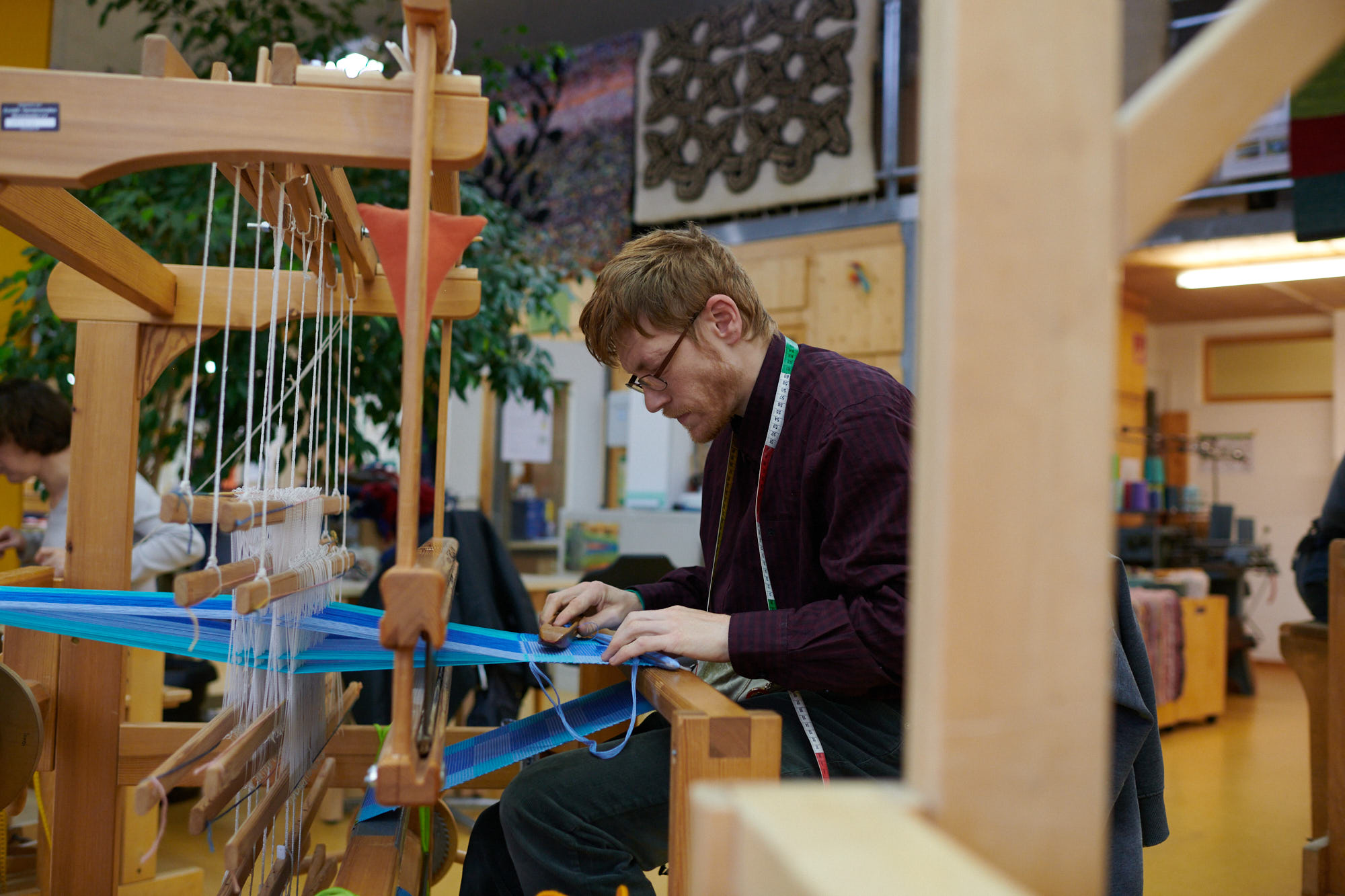 Mann arbeitet an einem traditionellen Webstuhl Ein Mann mit Brille und Maßband arbeitet konzentriert an einem traditionellen Webstuhl mit blauen Fäden.