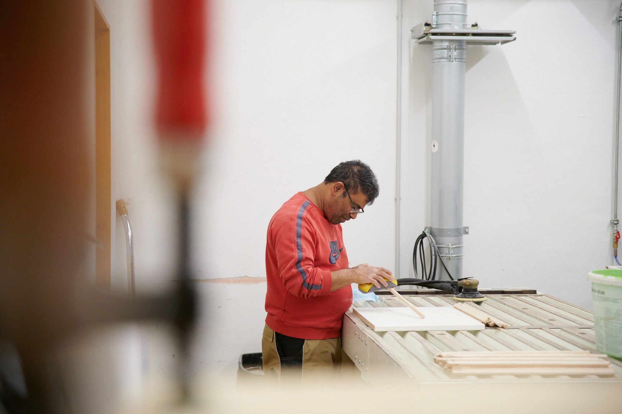 Handwerker beim Schleifen von Holzteilen in der Werkstatt Ein Handwerker in rotem Pullover schleift Holzteile an einem Tisch in einer Werkstatt.