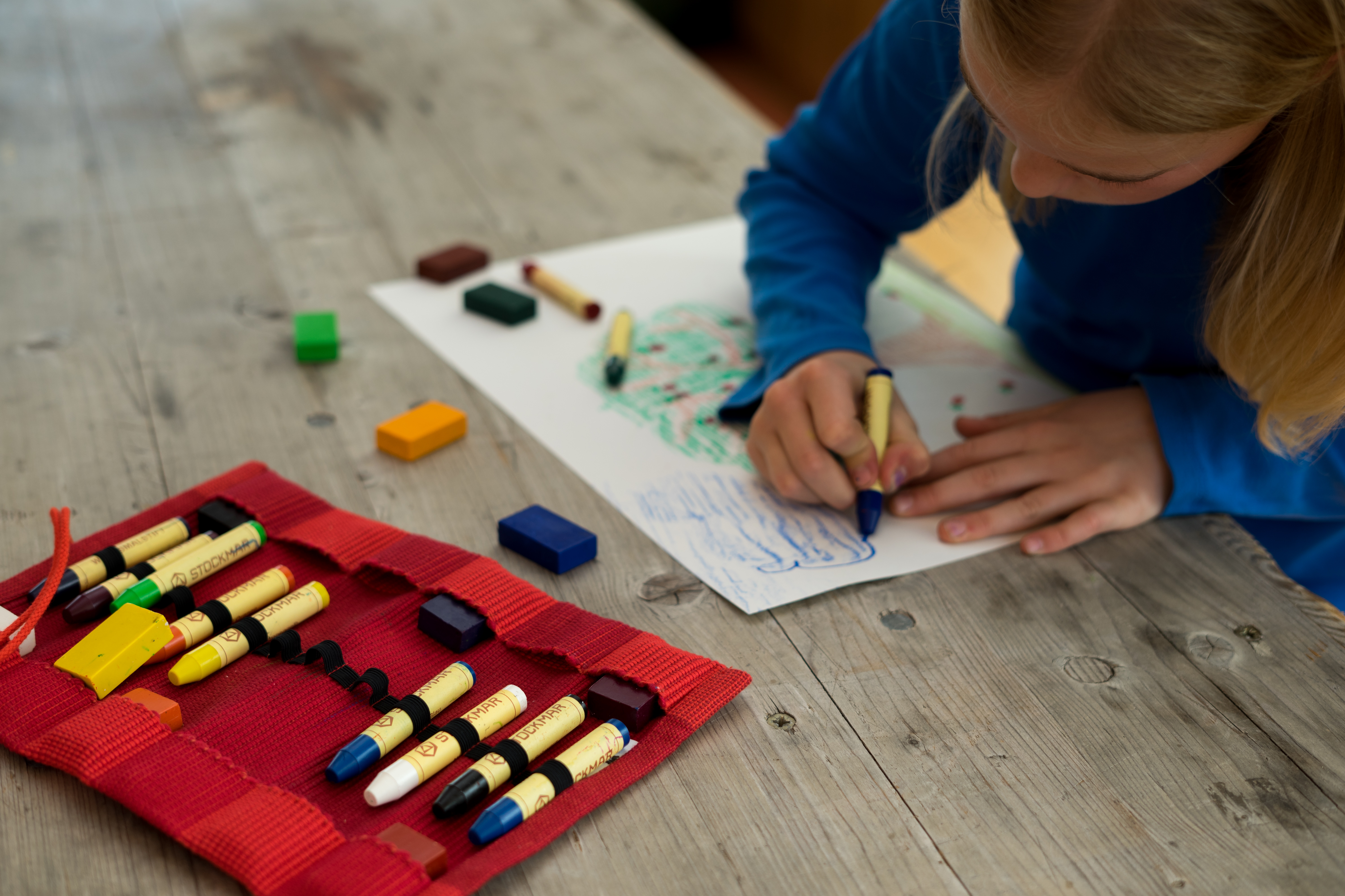 Kind malt mit Wachsmalstiften auf Papier Ein Kind zeichnet mit blauen Wachsmalstiften auf einem Blatt Papier. Daneben liegen weitere Stifte und ein rotes Etui auf dem Tisch.