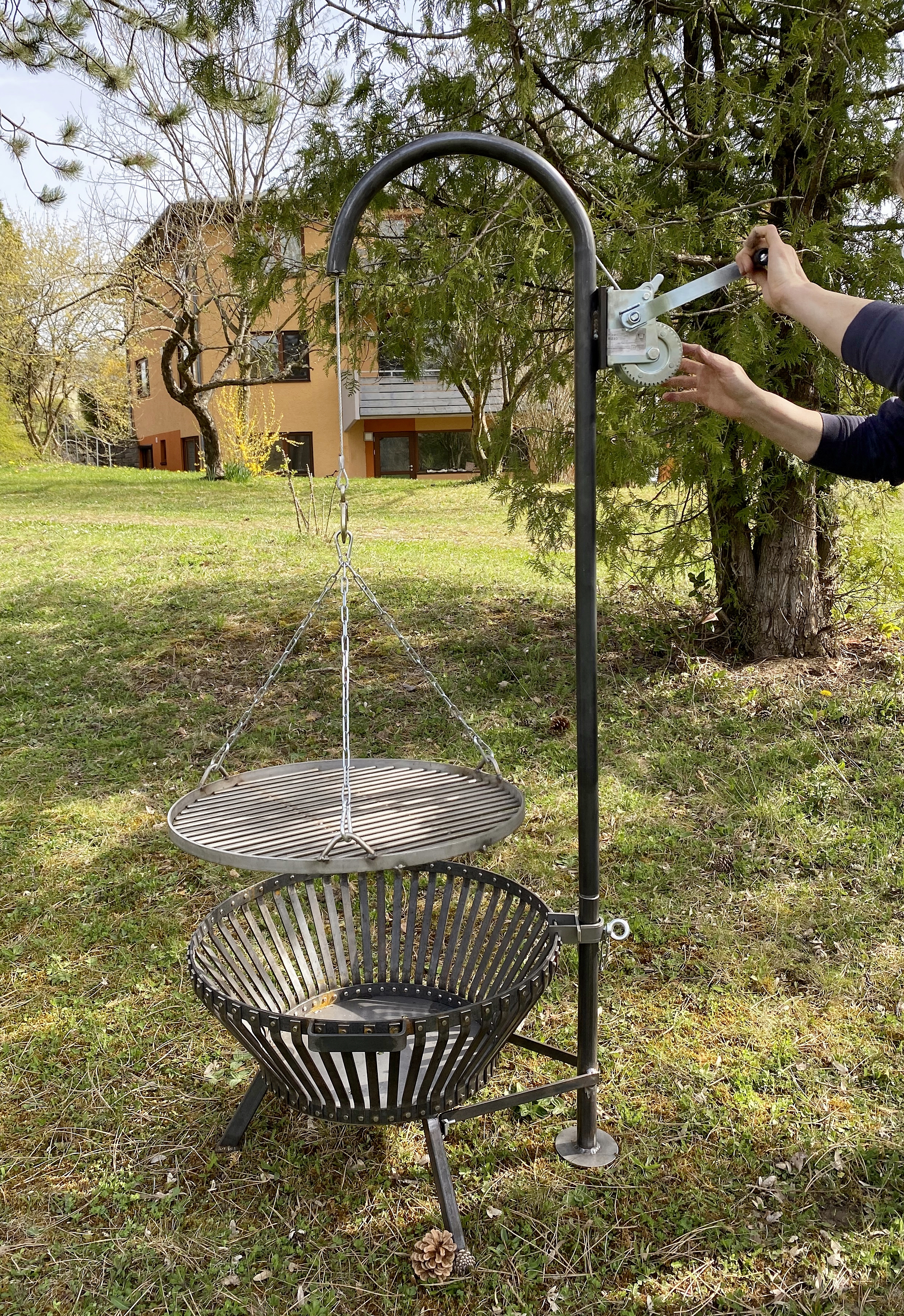 Schwarzer Schwenkgrill mit Feuerkorb, höhenverstellbarem Rost und Handkurbel auf einer Wiese im Garten