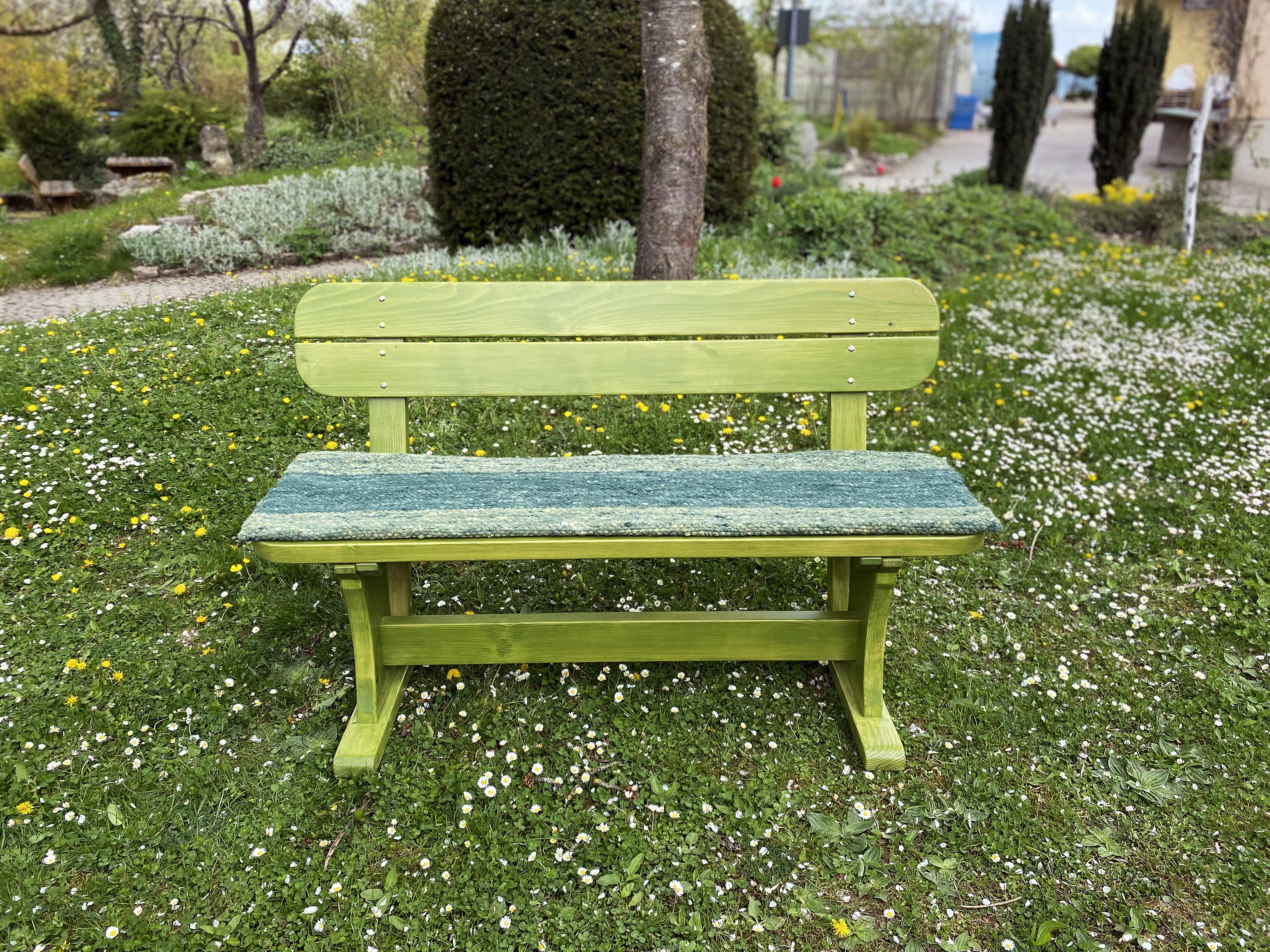 Grün lasierte Holz-Gartenbank mit Rückenlehne und blauem Sitzpolster auf einer Wiese voller Gänseblümchen im Garten