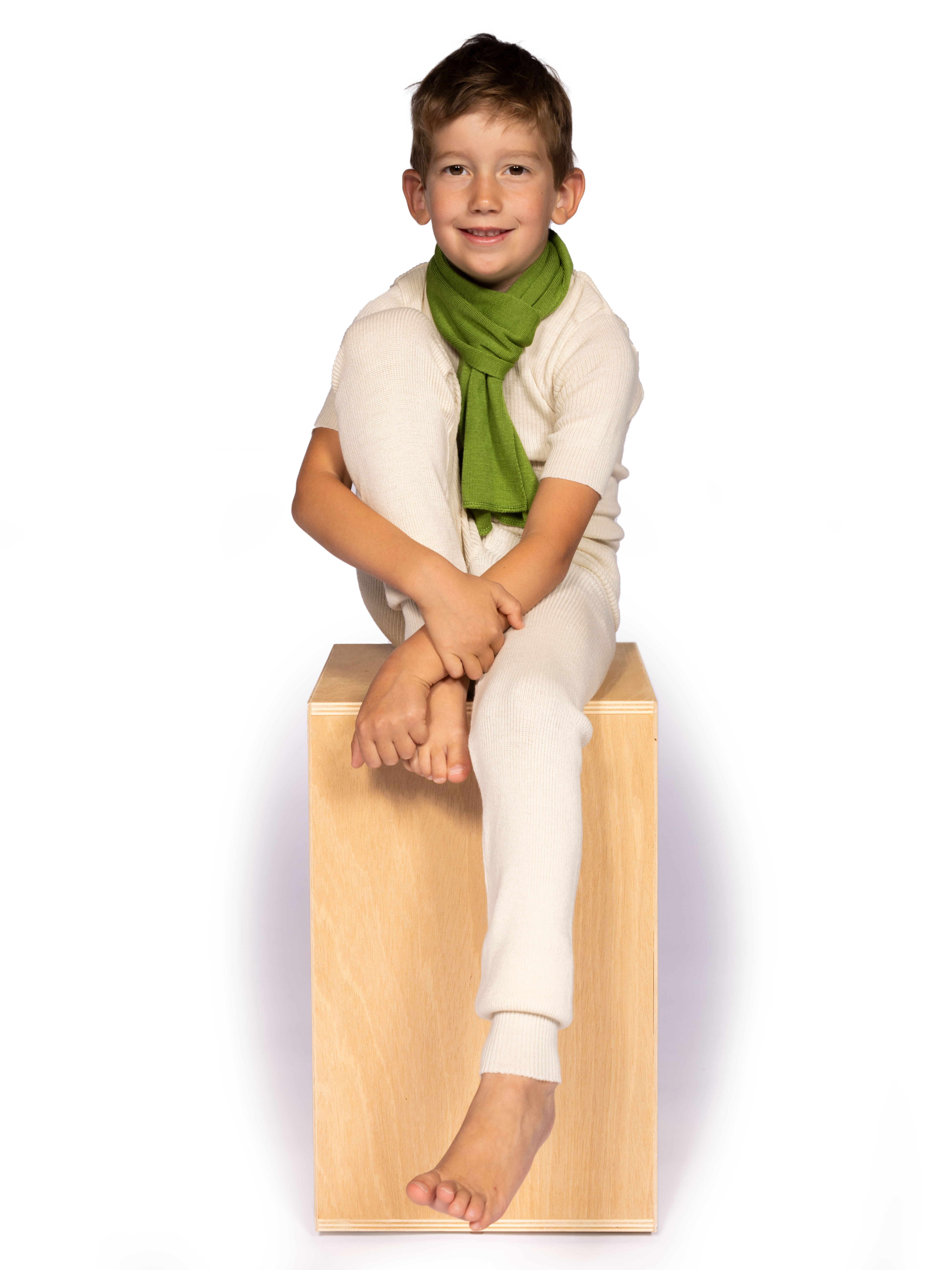 Lächelnder Junge mit grünem Schal und weißem Outfit, der auf einer Holzkiste sitzt