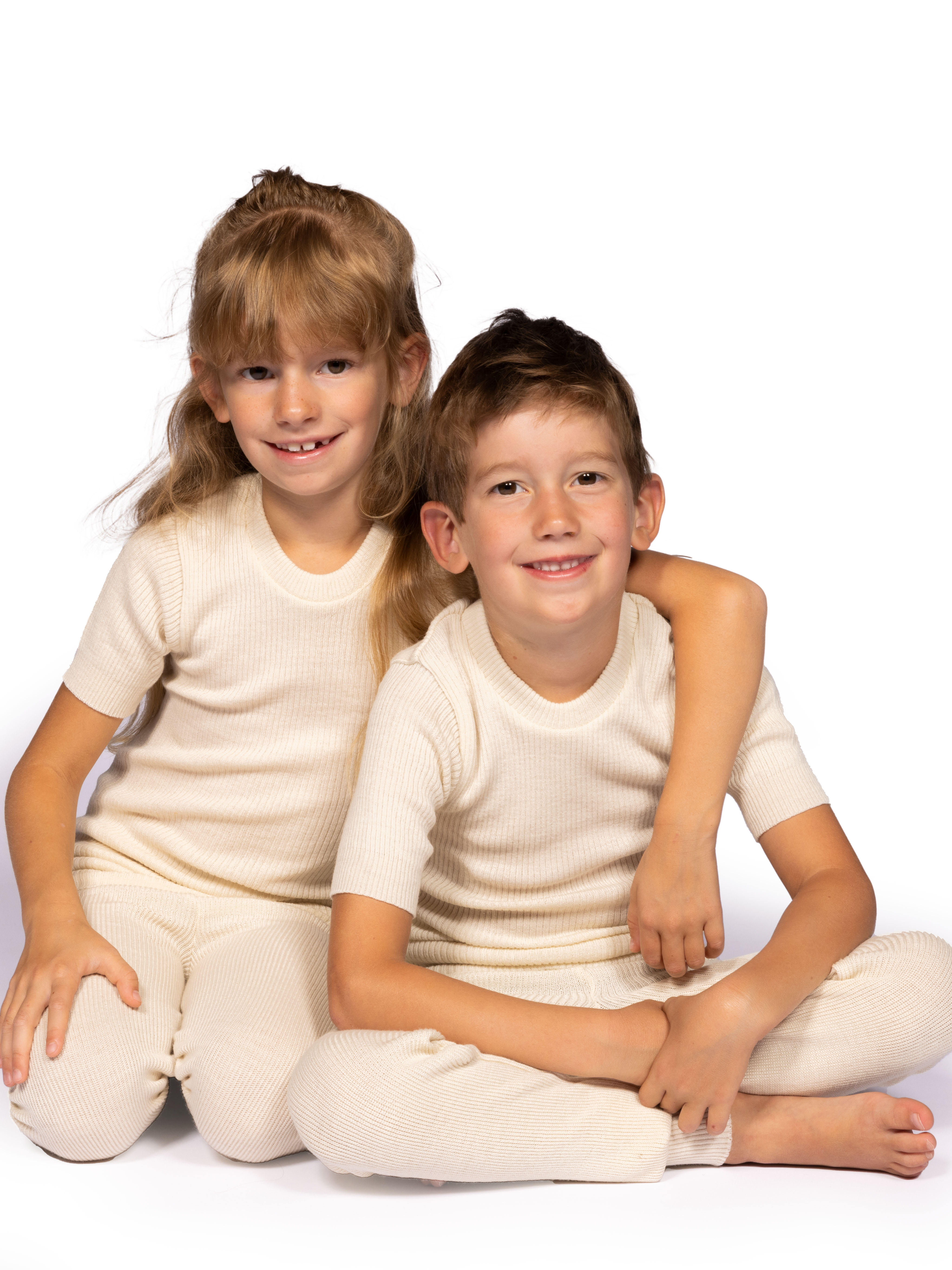 Zwei Kinder in beiger, gerippter Freizeitkleidung, lachend und nah beieinander sitzend