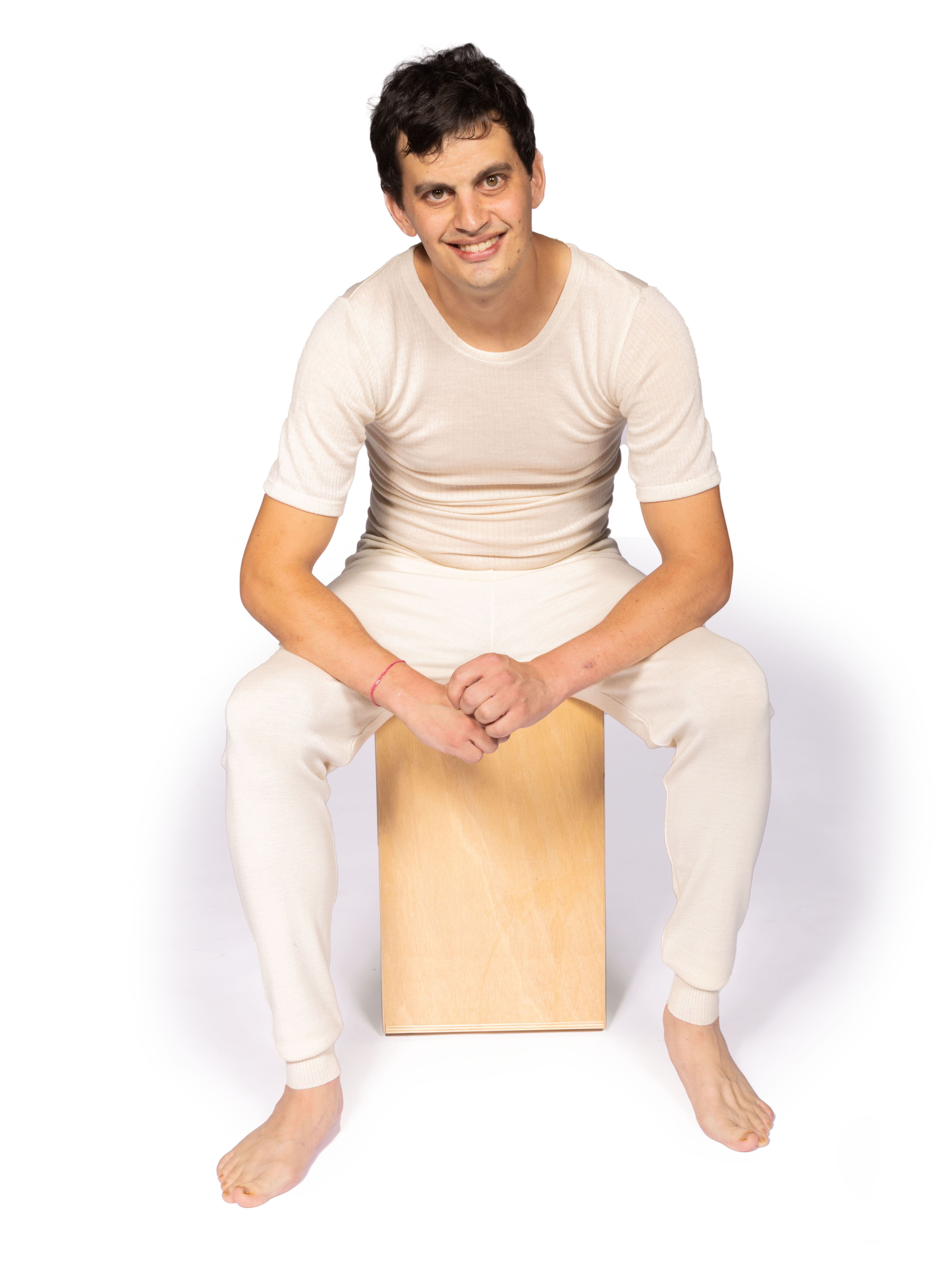 Mann in bequemer, cremefarbener Loungewear sitzt auf Holzschemel Ein lächelnder Mann in cremefarbener Loungewear sitzt barfuß auf einem Holzschemel vor weißem Hintergrund.