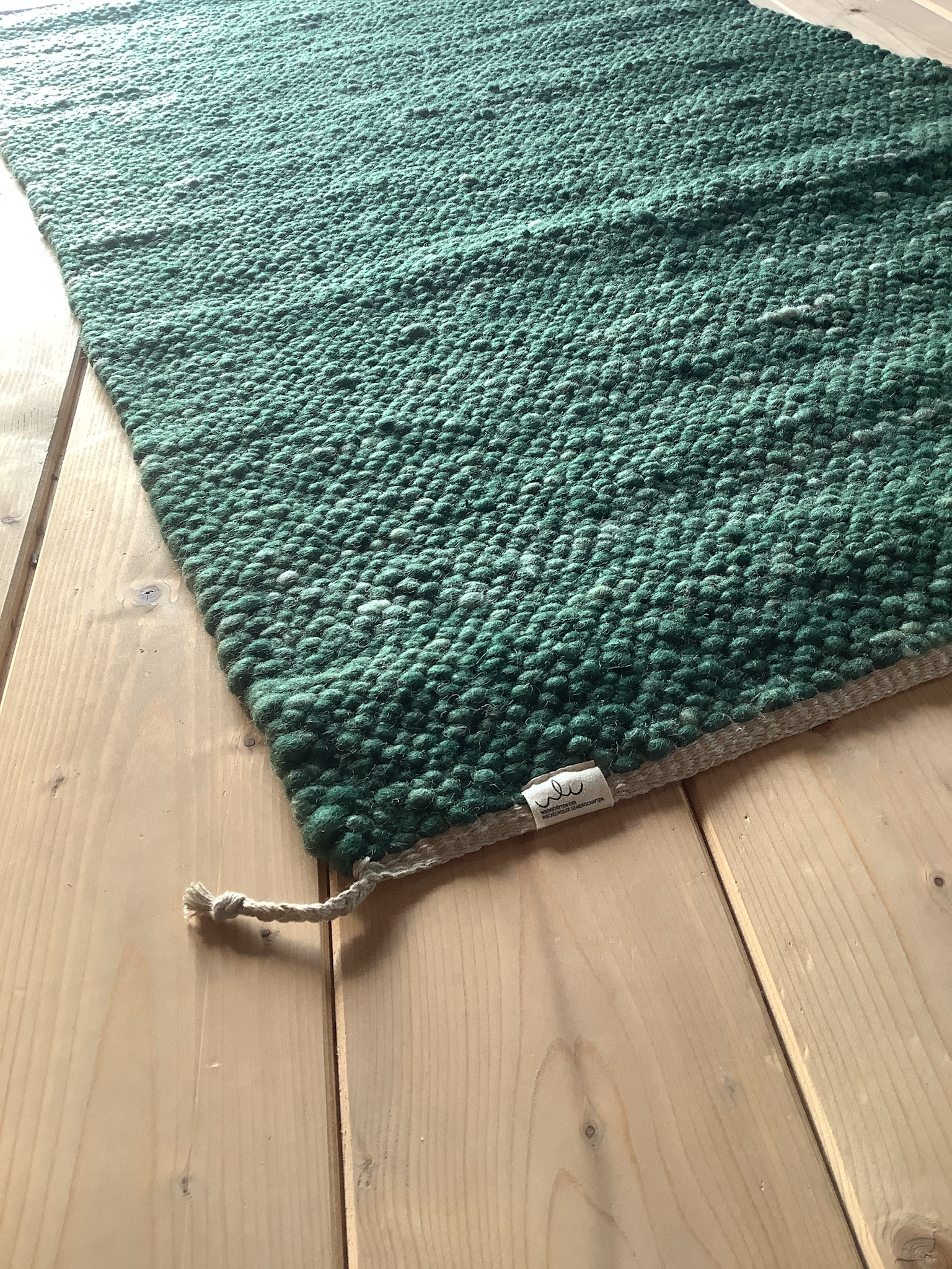 Nahaufnahme eines handgewebten grünen Wollteppichs mit dicker Struktur auf hellem Holzboden, sichtbarem Etikett und Kordel am Rand
