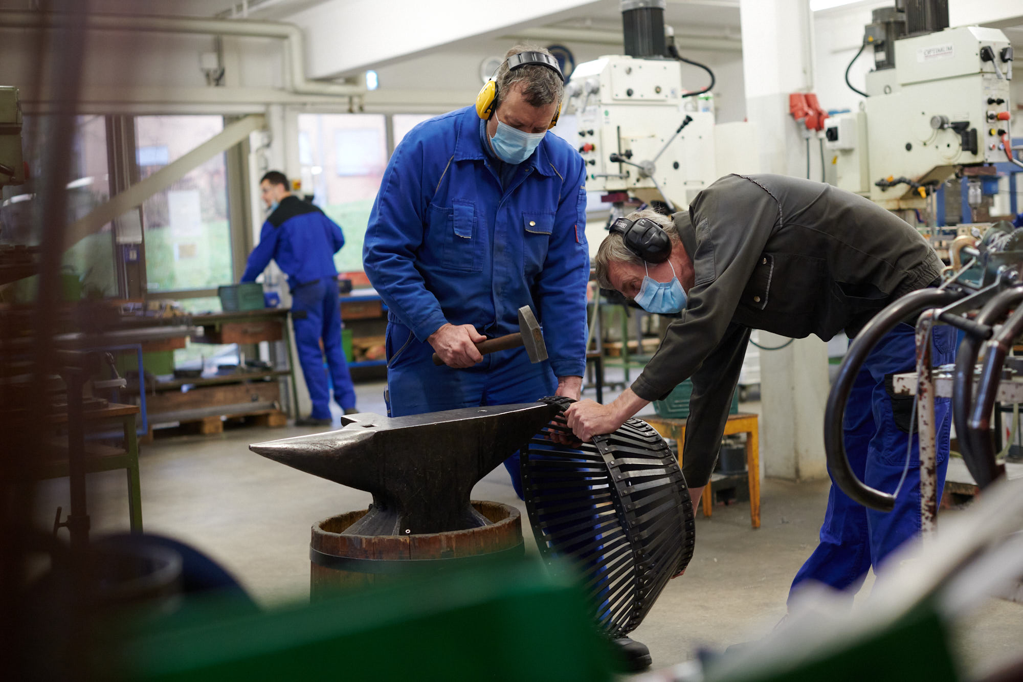 Zwei Fachkräfte in Schutzkleidung bearbeiten in einer Metallwerkstatt ein Bauteil am Amboss mit Hammer und Gehörschutz.