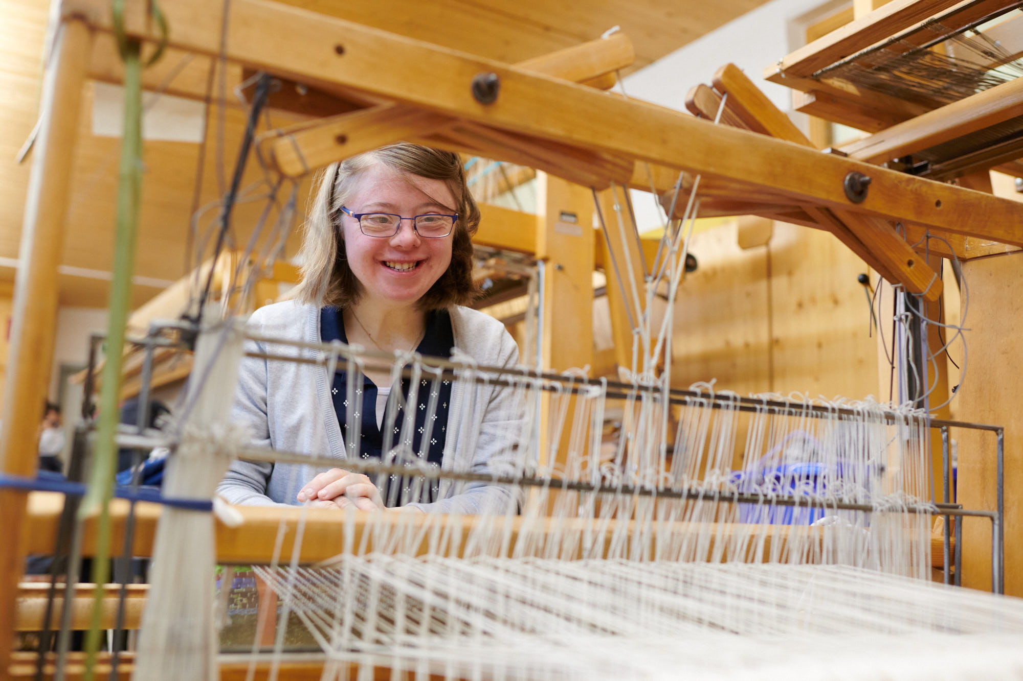 Lächelnde Frau an einem Webstuhl in einer Werkstatt Junge Frau mit Brille arbeitet fröhlich an einem traditionellen Webstuhl in einer hellen Werkstatt