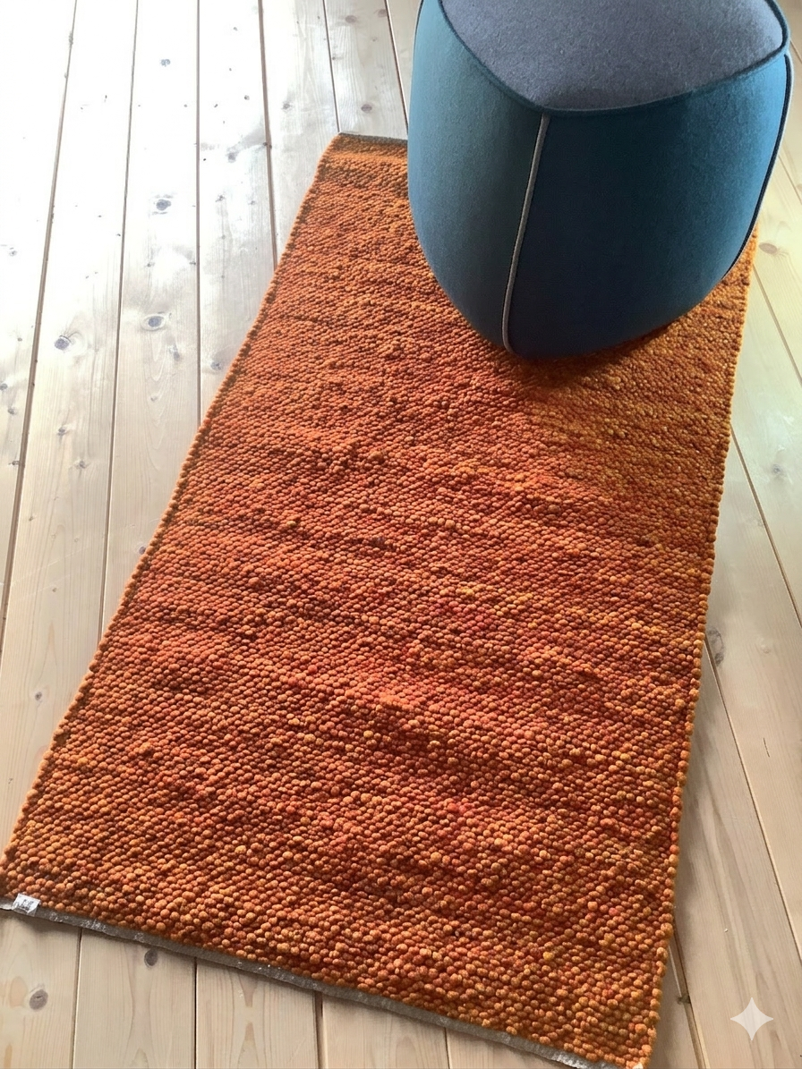Rechteckiger, orange melierter Wollteppich mit grober, kugeliger Struktur auf hellem Holzboden, dekoriert mit einem modernen Hocker