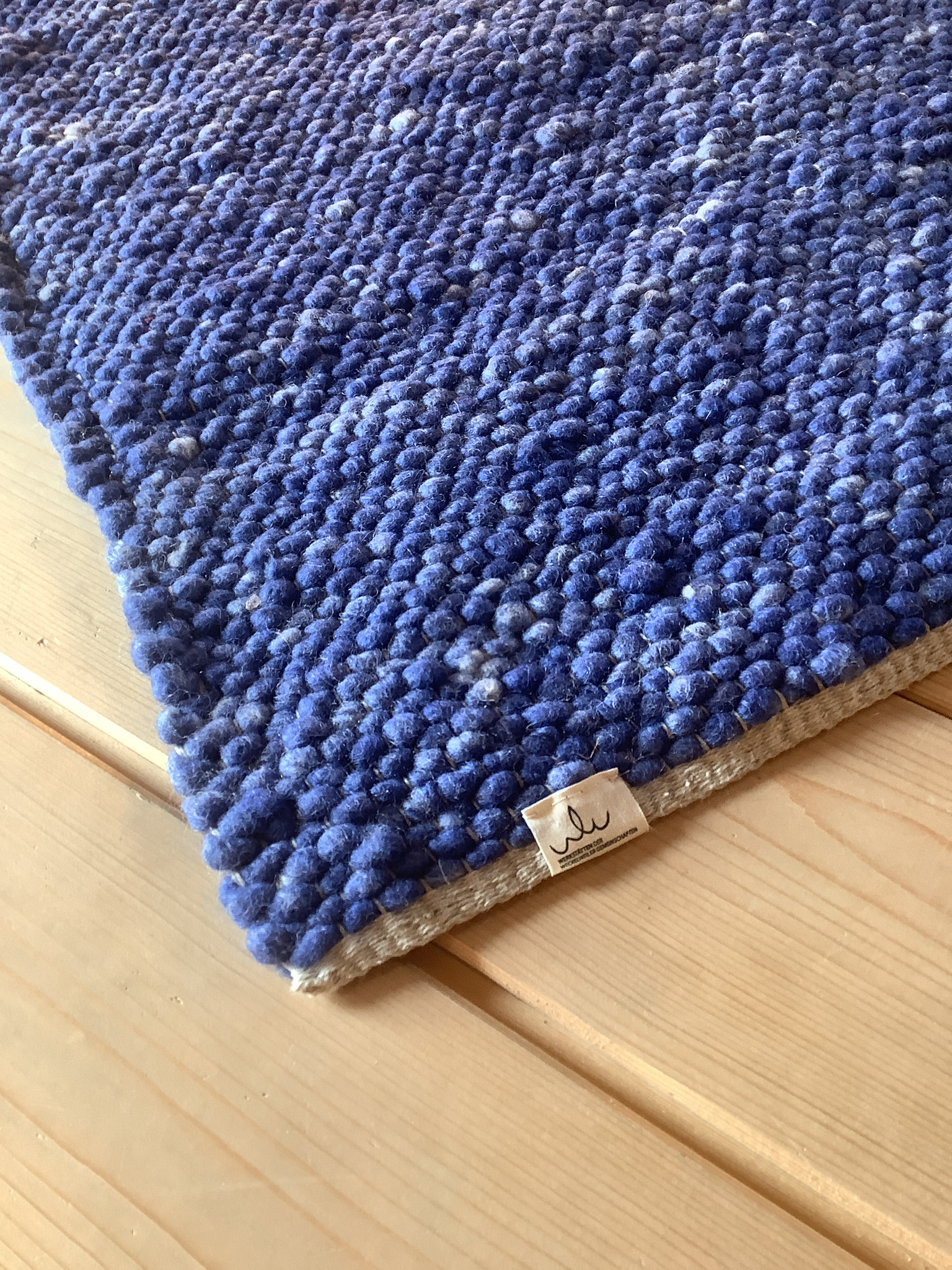 Detailansicht eines blauen, grob strukturierten Wollteppichs mit Labelkante auf hellem Holzfußboden