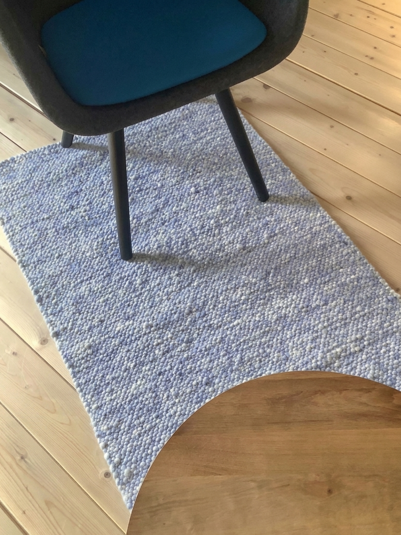 Nahaufnahme eines modernen Bürostuhls mit blauem Sitzpolster auf einem hellblauen strukturierten Teppichläufer auf hellem Holzboden, teilweise mit rundem Holztisch im Vordergrund