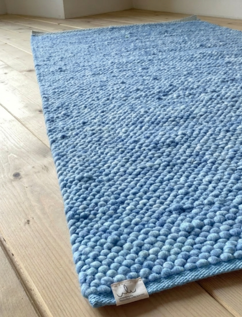 Nahaufnahme eines handgewebten hellblauen Wollteppichs mit grober Struktur, rechteckig auf hellem Holzboden liegend
