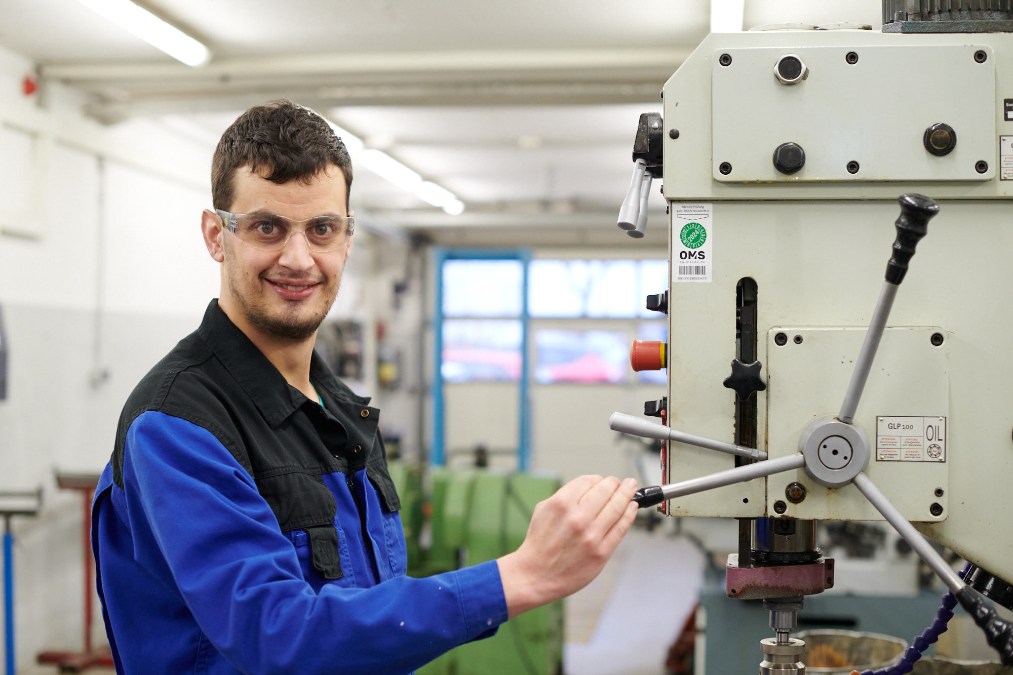 Facharbeiter in blauer Arbeitskleidung mit Schutzbrille an einer Ständerbohrmaschine in der Metallwerkstatt