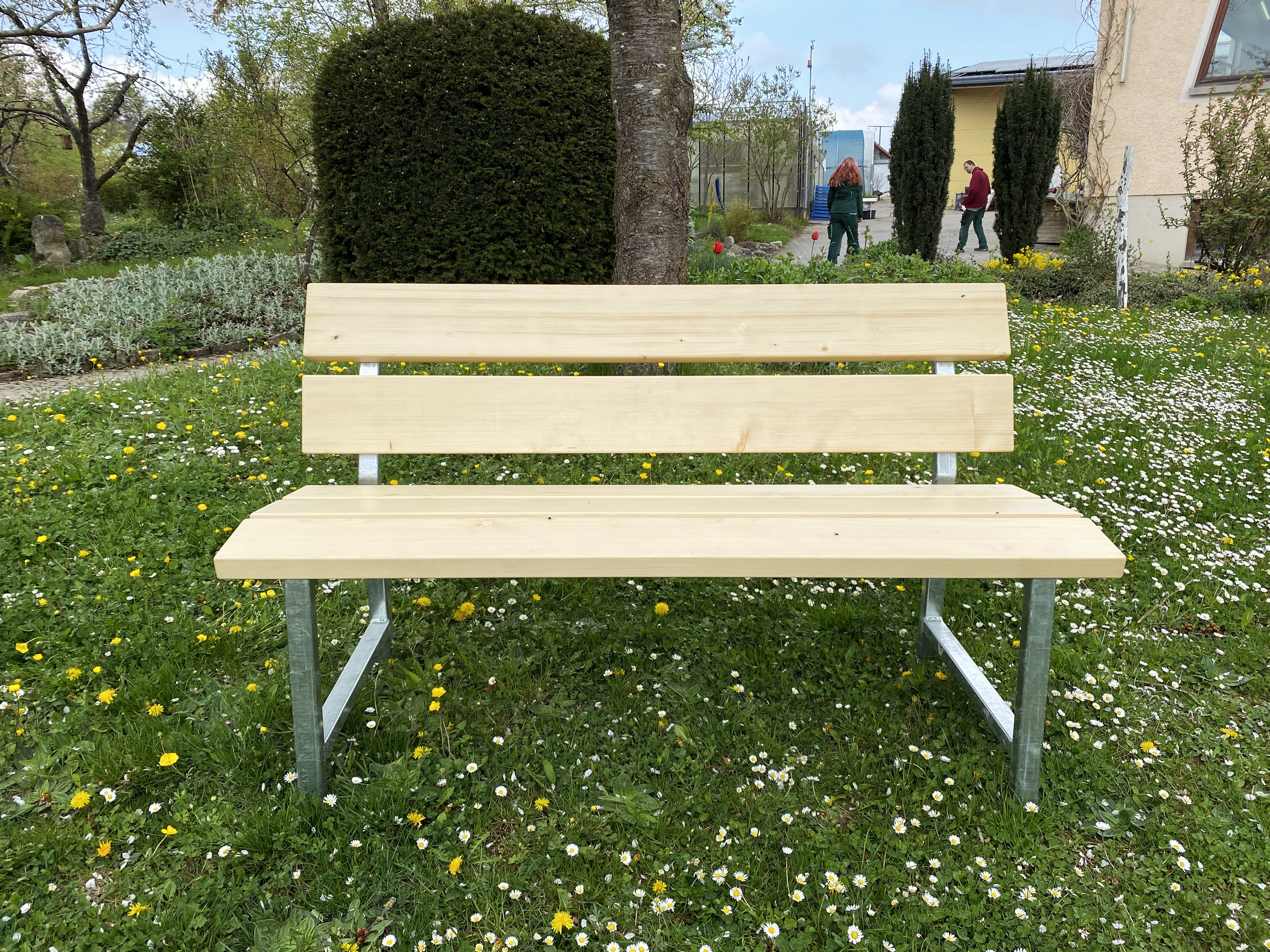 Helle Gartenbank aus Holz mit Rückenlehne und verzinktem Metallgestell auf einer Wiese im Garten